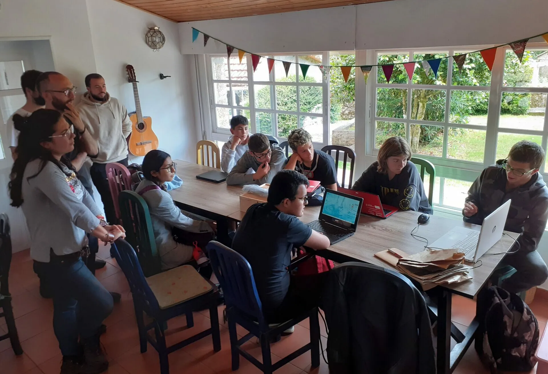 Xente traballando con portátiles e papeis arredor dunha mesa longa nunha sala luminosa con bandeiriñas e unha guitarra na parede