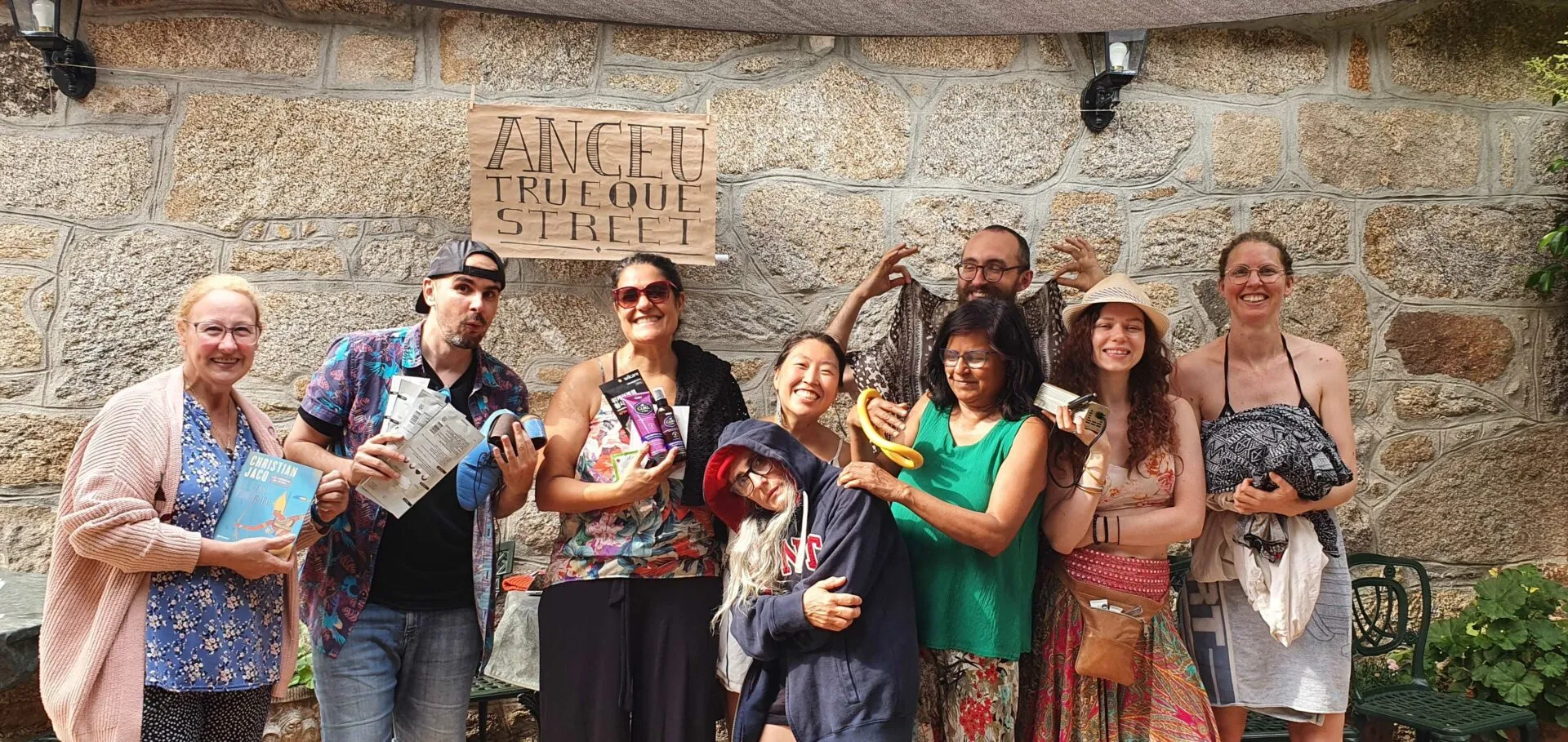Nueve personas alegres con ropa de colores posando frente a un muro de piedra bajo un cartel de madera, con folletos y pequeños objetos en las manos