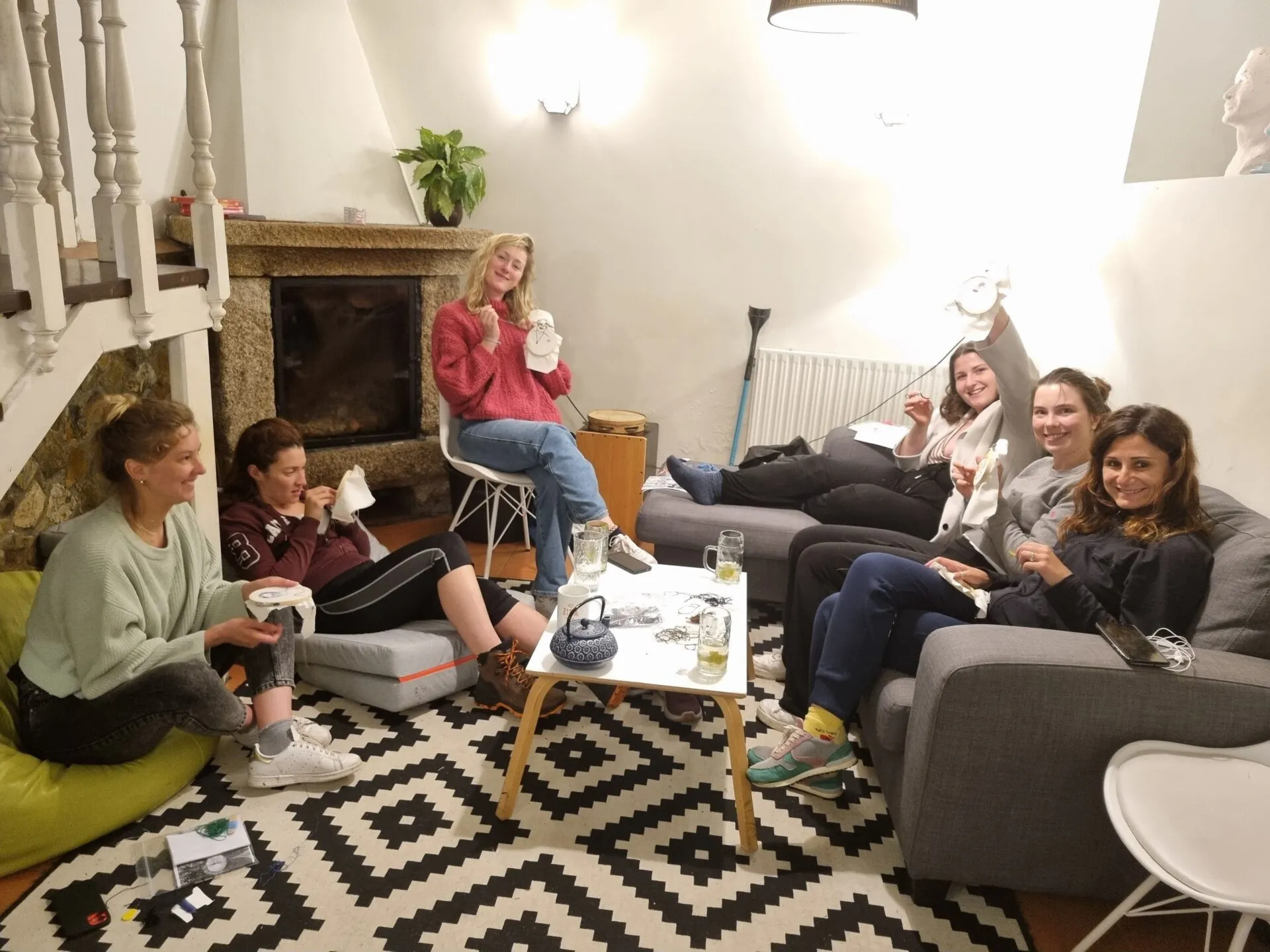 Seis mulleres relaxadas en sofás e coxíns onda unha lareira de pedra, suxeitando bastidores de bordado nun salón acolledor