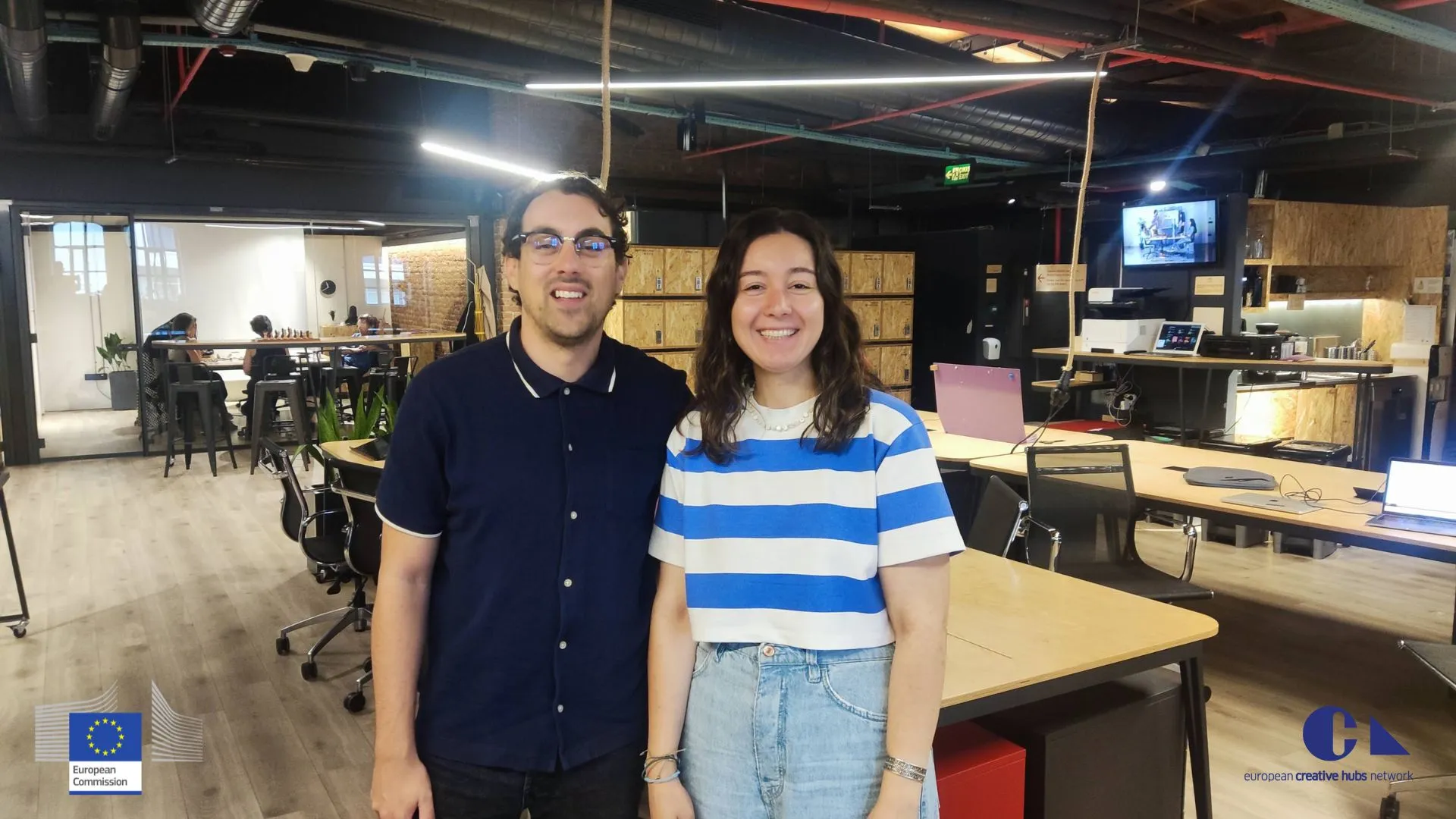 Un home e unha muller sorrindo xuntos nun espazo de coworking moderno con mesas de madeira e o logotipo da Comisión Europea nunha esquina