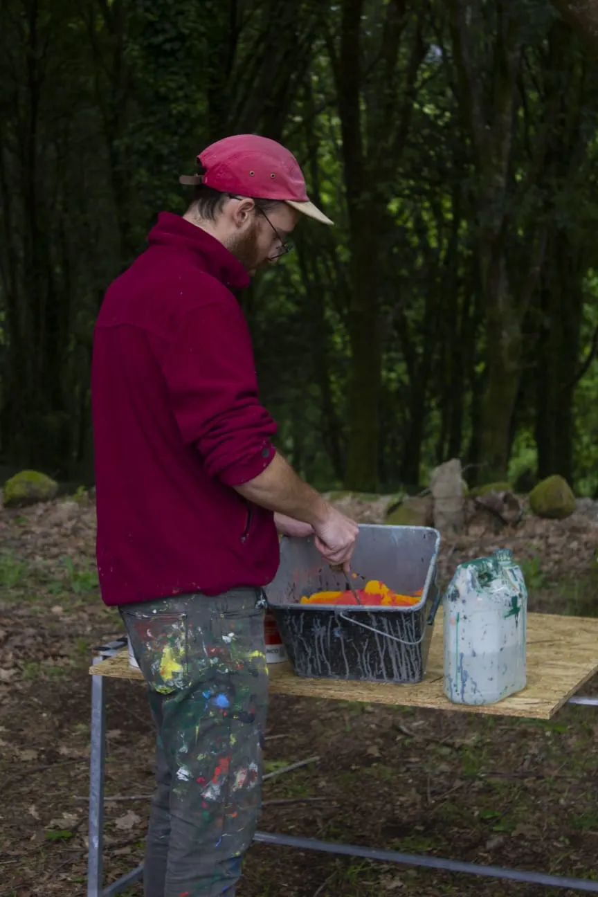 Home con gorra vermella e pantalón salpicado de pintura remexendo pintura laranxa nun caldeiro sobre unha mesa no bosque