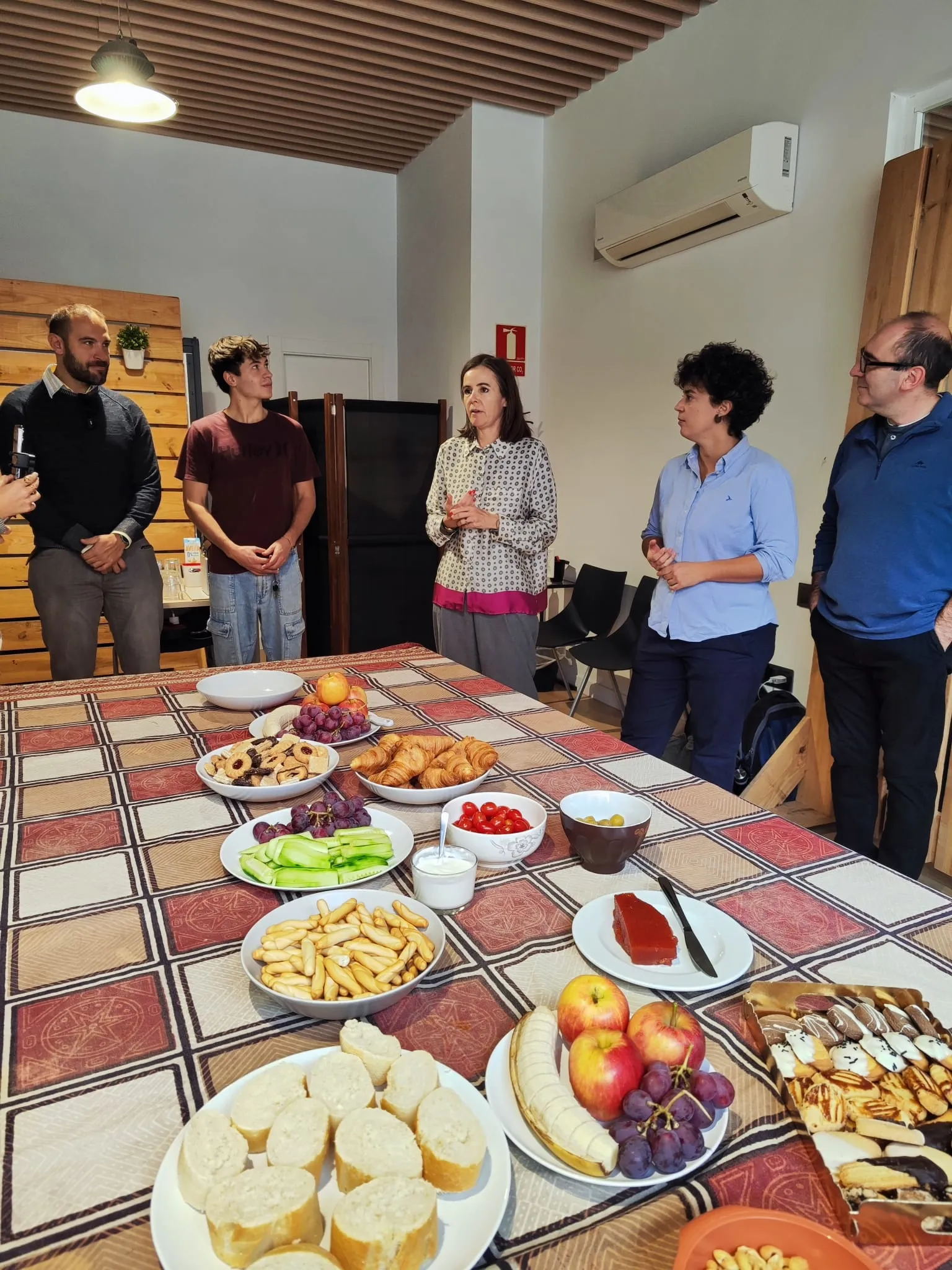 Cinco personas de pie alrededor de una mesa larga con fruta, cruasanes, colines y dulces mientras una mujer habla