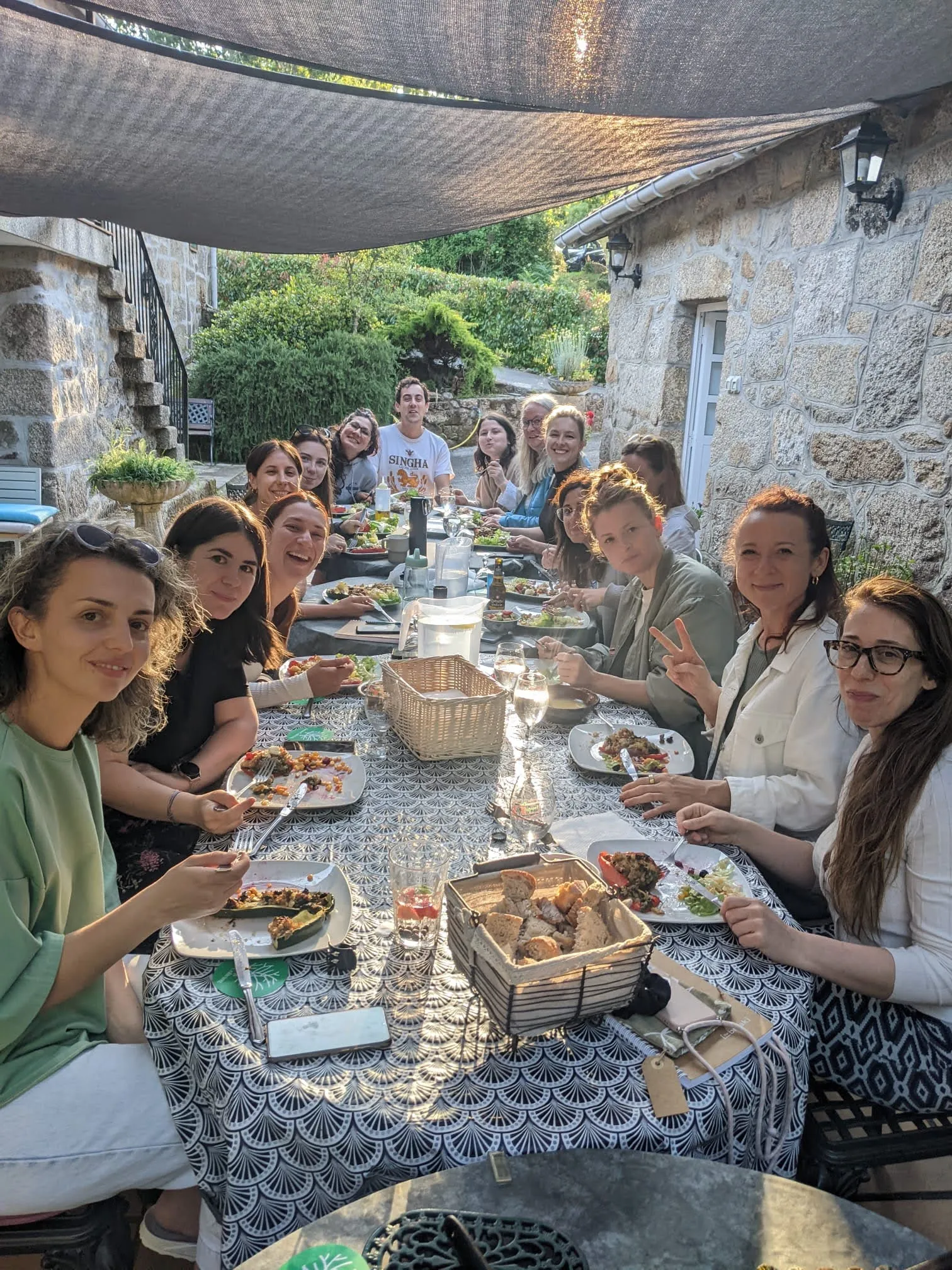 Mesa longa con sobre todo mulleres comendo verduras recheas baixo unha vela de sombra a carón dunha casa de pedra