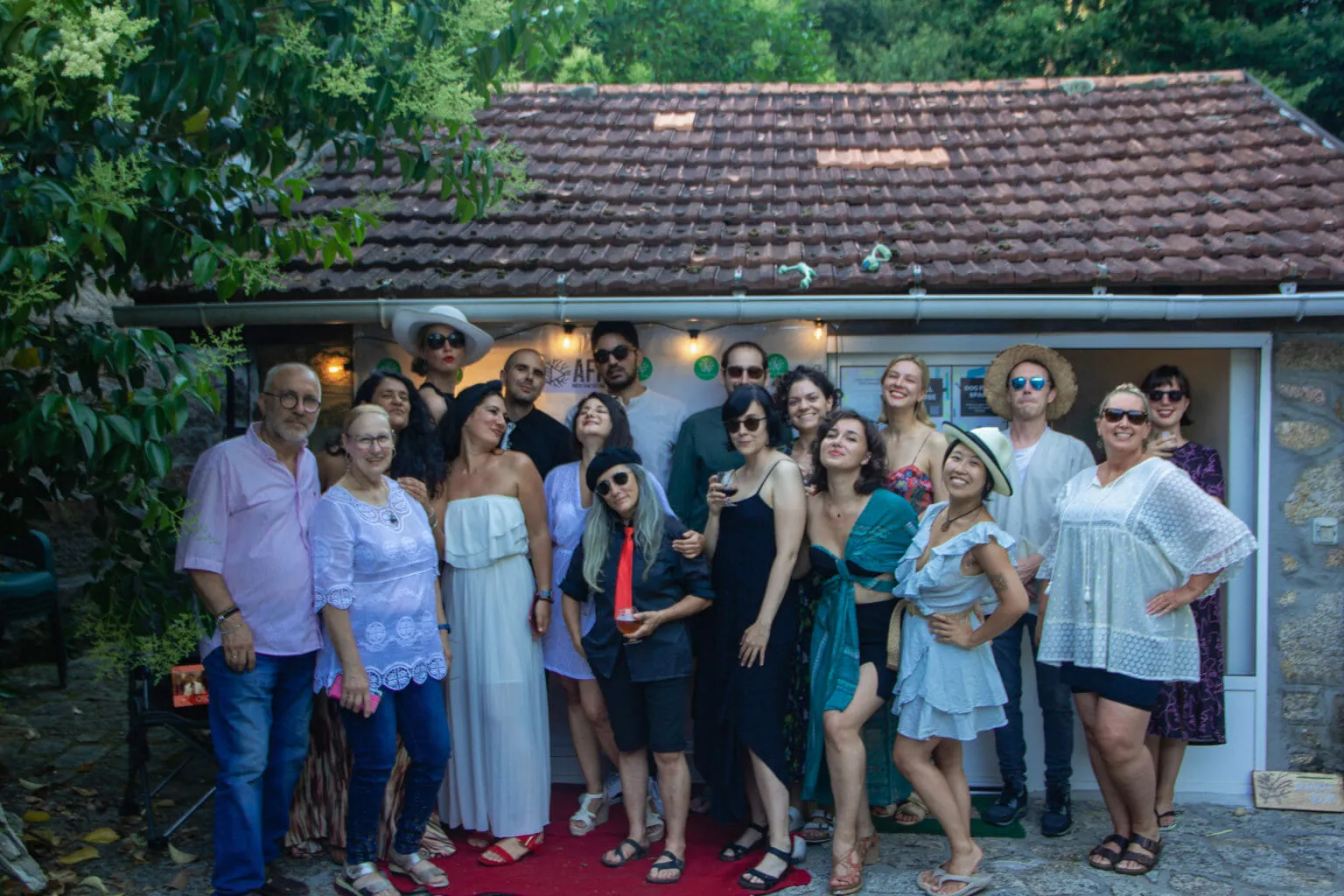 Unhas vinte persoas arranxadas posando diante dunha pequena casa de pedra cunha alfombra vermella na porta