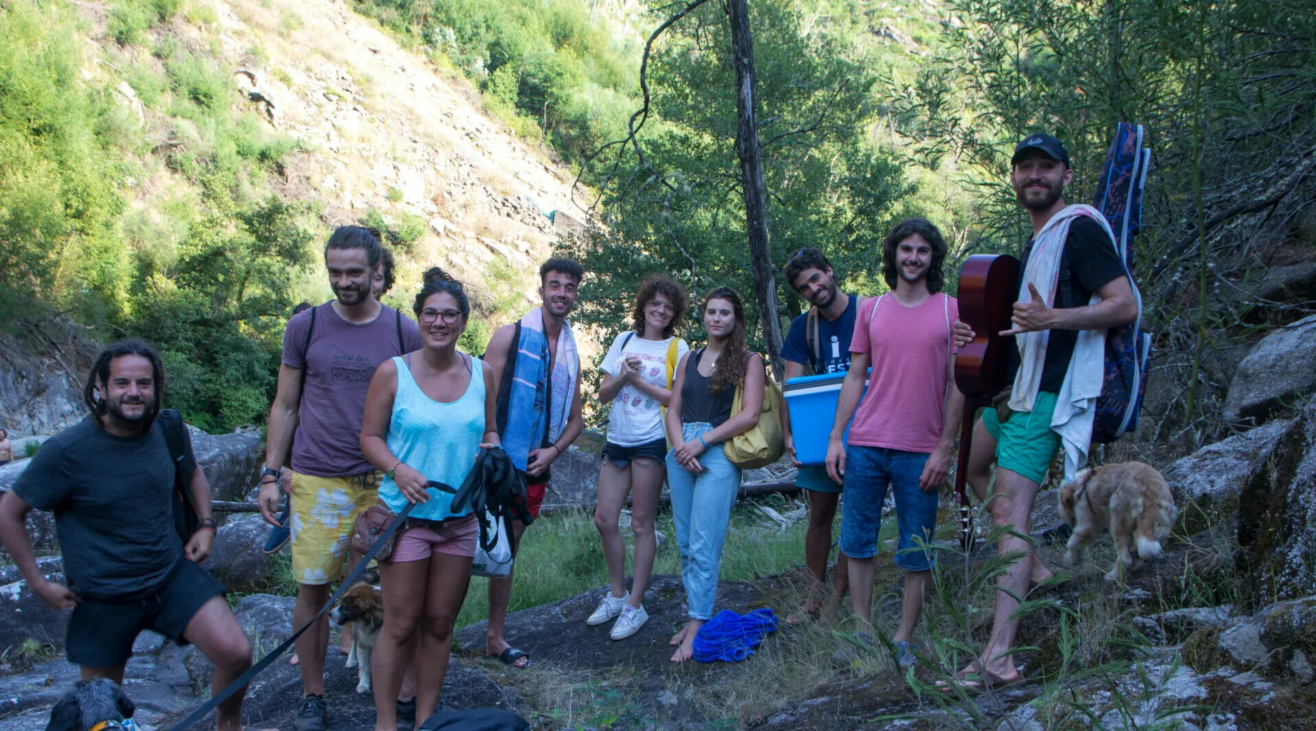 Diez personas con toallas, una guitarra y una nevera portátil posando junto a un arroyo rocoso en una garganta boscosa, con un perro a la derecha