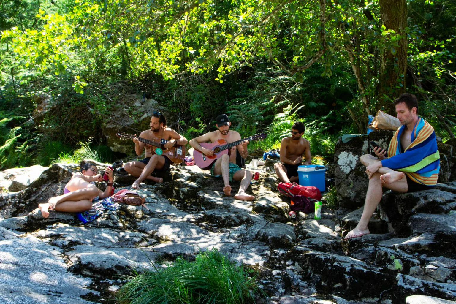 Gente relajándose sobre rocas soleadas junto a un arroyo, dos de ellos tocando la guitarra
