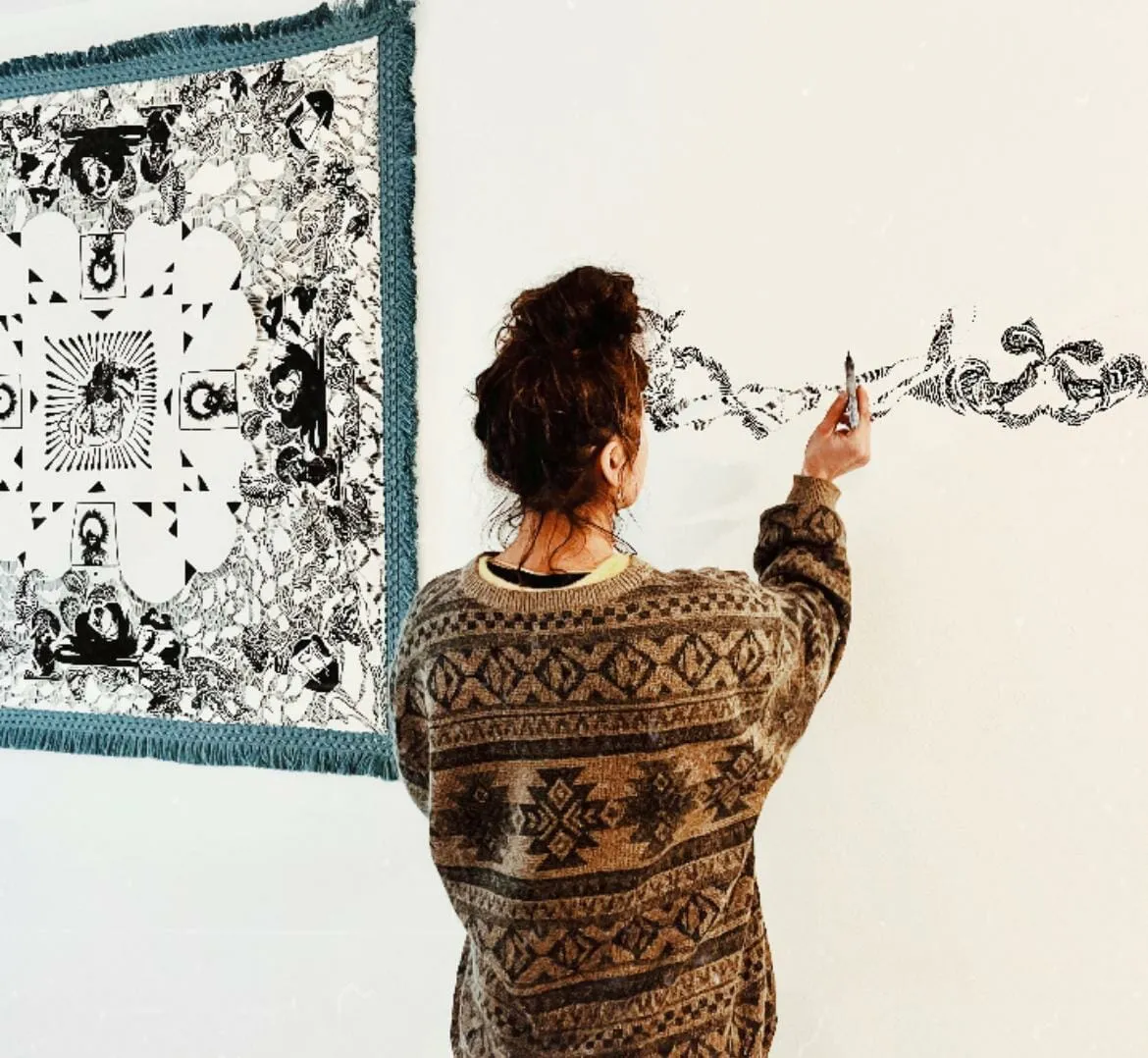 Mujer de espaldas dibujando un intrincado motivo negro directamente sobre una pared blanca, junto a un tapiz con flecos de arte similar
