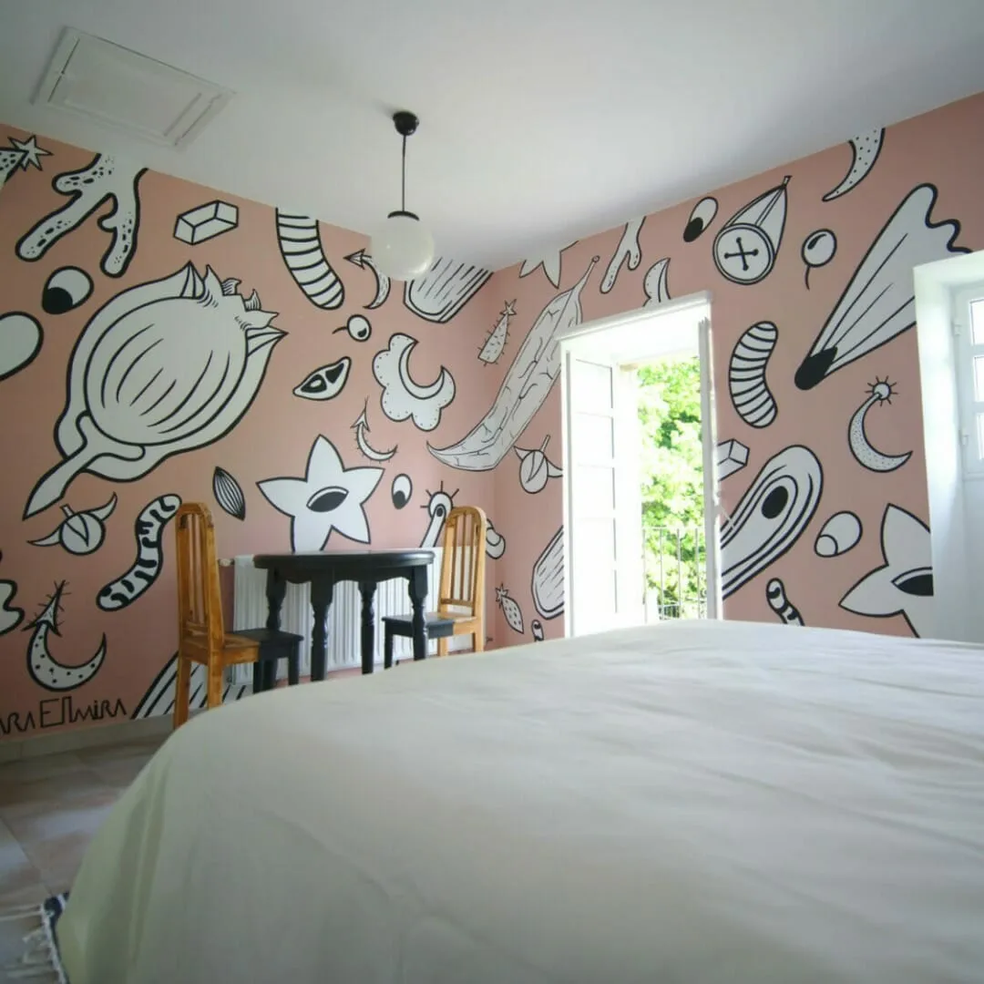 Habitación con paredes rosas cubiertas de murales de garabatos en blanco y negro, una cama blanca amplia y una puerta abierta al verde
