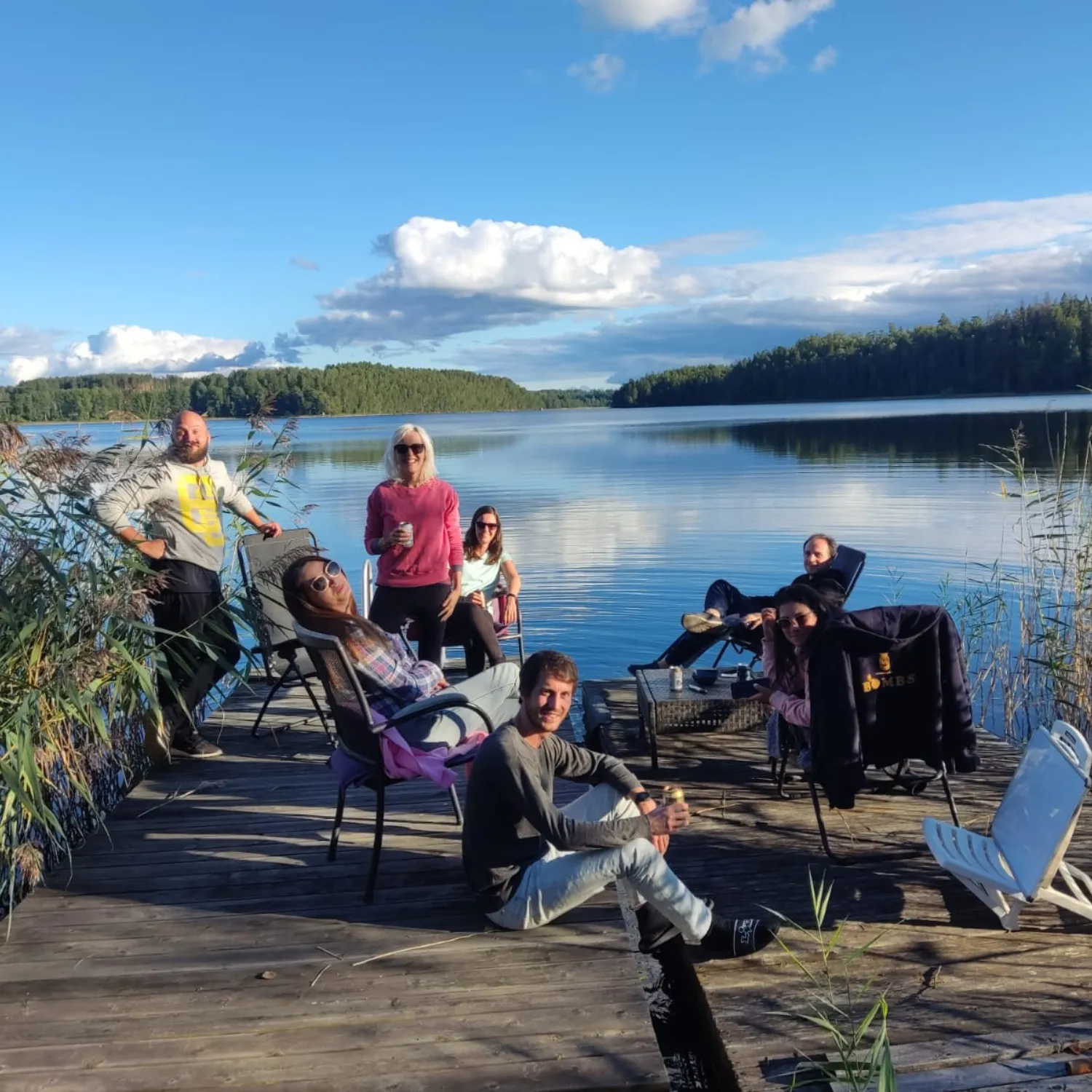 Sete persoas descansando en cadeiras pregables nun pantalán de madeira a carón dun lago tranquilo rodeado de bosque, baixo un ceo azul