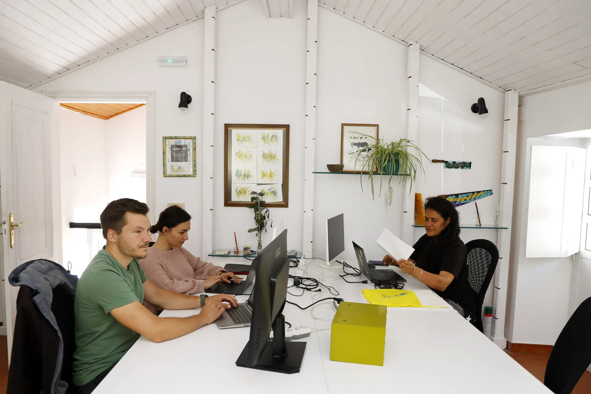 Tres persoas traballando con portátiles nunha longa mesa branca nunha sala de coworking luminosa con teito de madeira a dúas augas