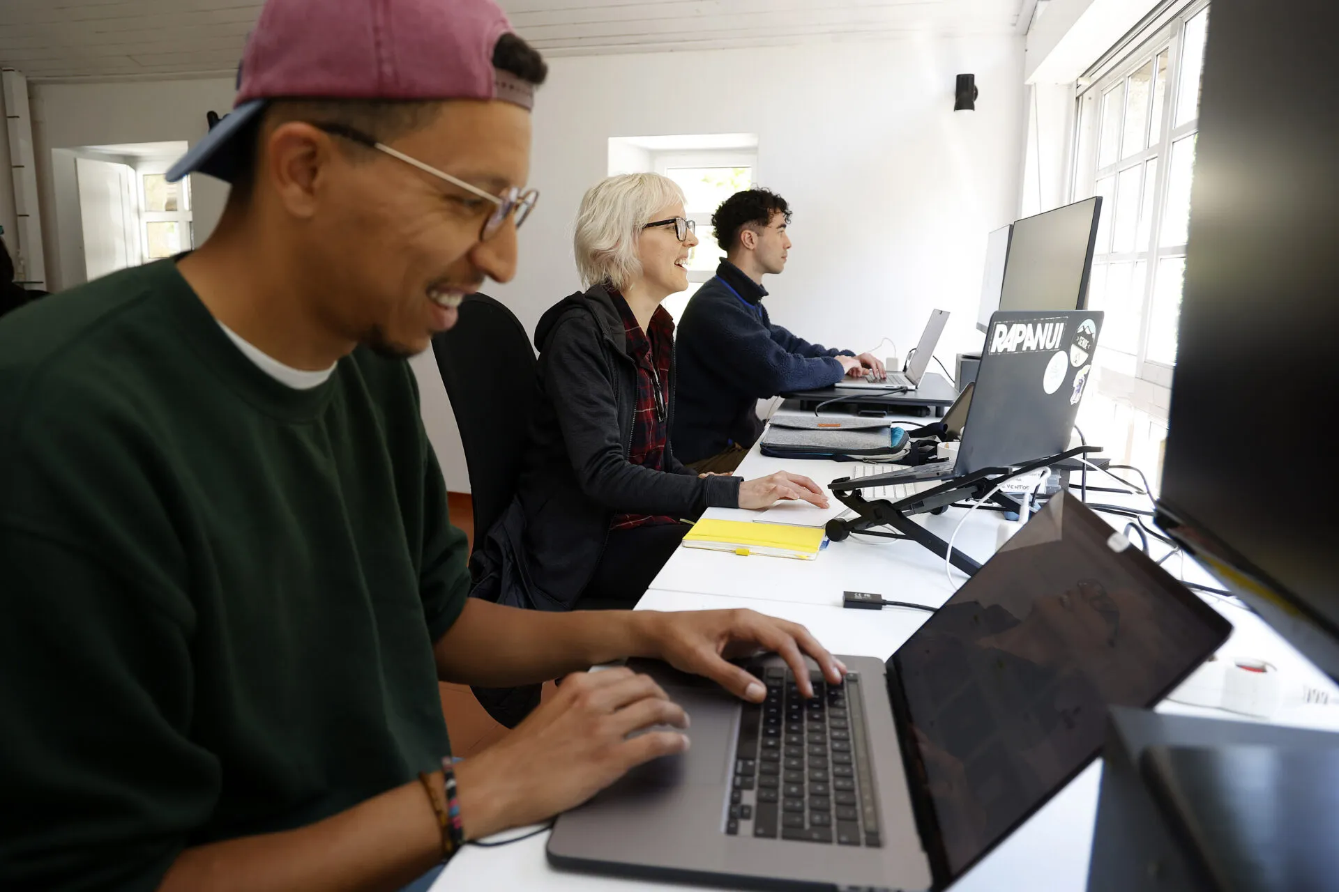 Tres persoas escriben nos seus portátiles ao longo dunha mesa branca nunha sala de coworking luminosa
