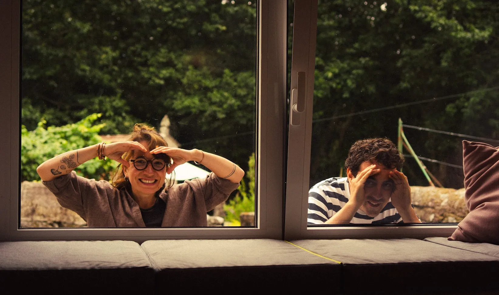 Dos personas asomándose por la ventana desde el jardín, con las manos enmarcando la cara, vistas desde un banco con cojines