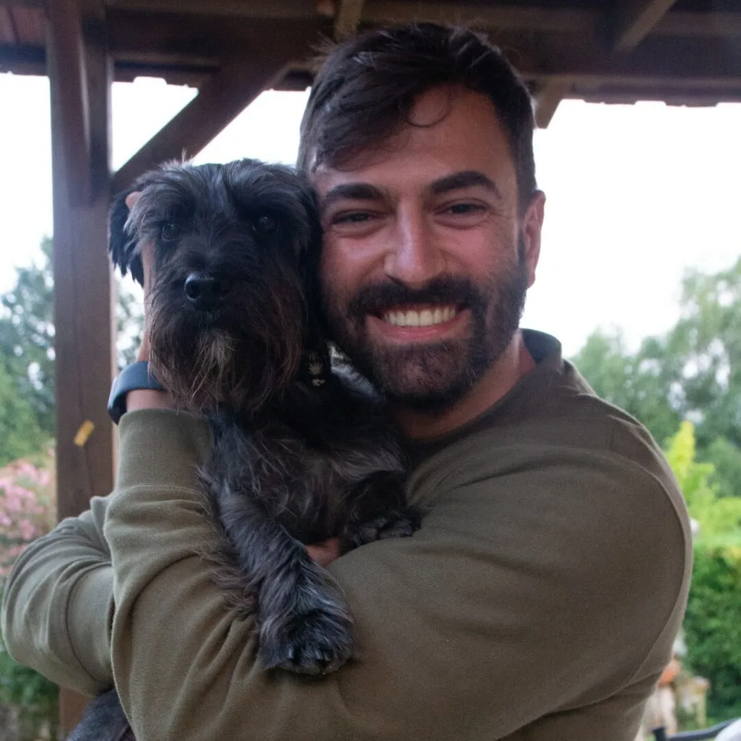 Hombre barbudo sonriente sosteniendo en brazos a un perro pequeño de pelo negro y duro bajo un porche de madera