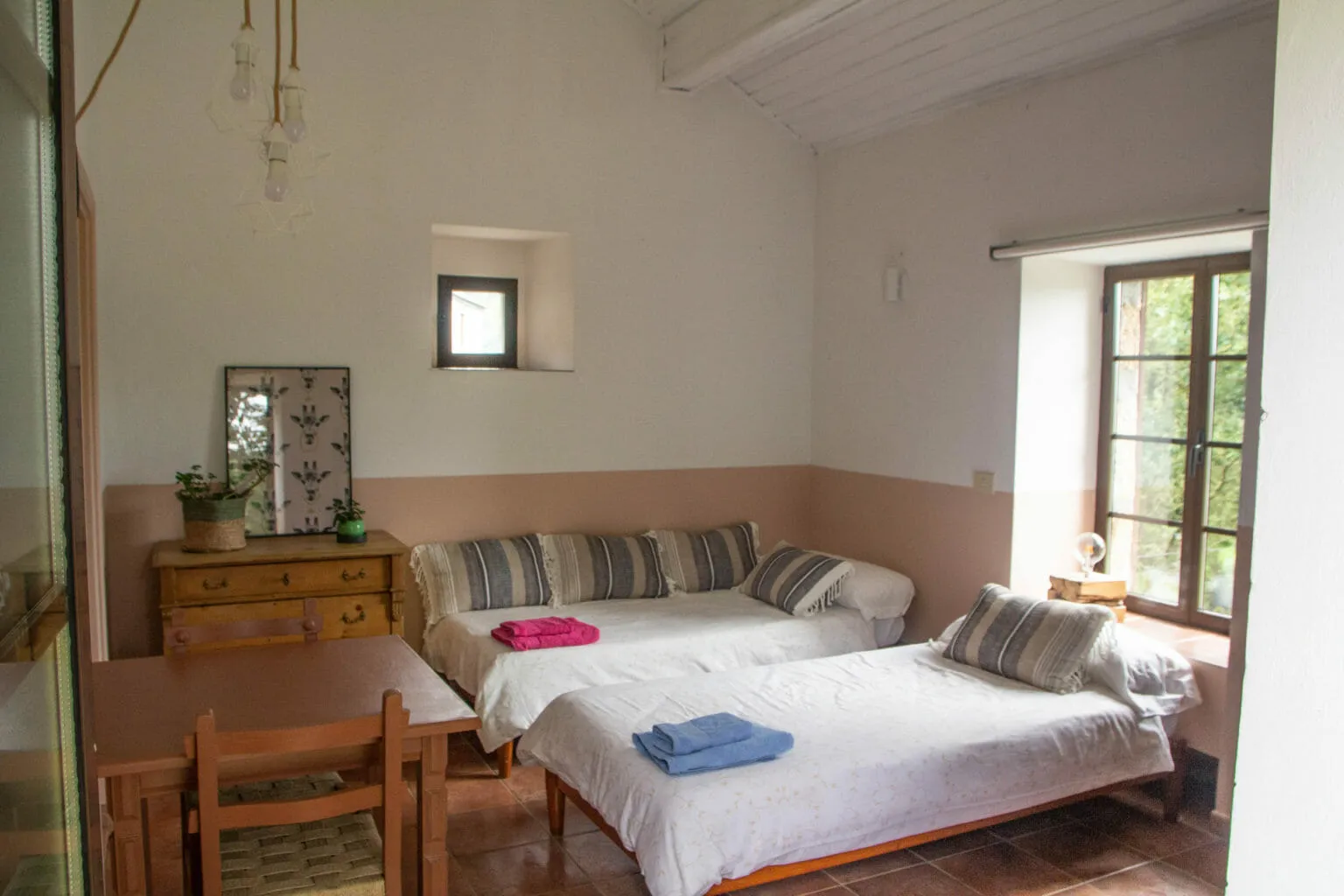 Una habitación luminosa de paredes blancas con dos camas individuales con cojines de rayas, una cómoda de madera y una ventana con contraventanas
