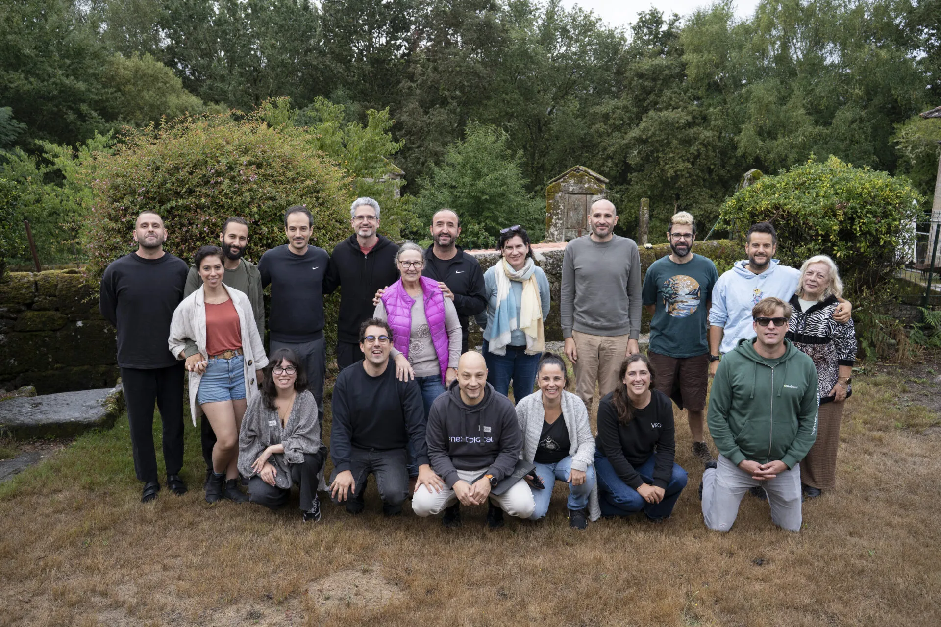 Una veintena de personas posando juntas sobre hierba seca delante de muros de piedra antiguos y vegetación densa