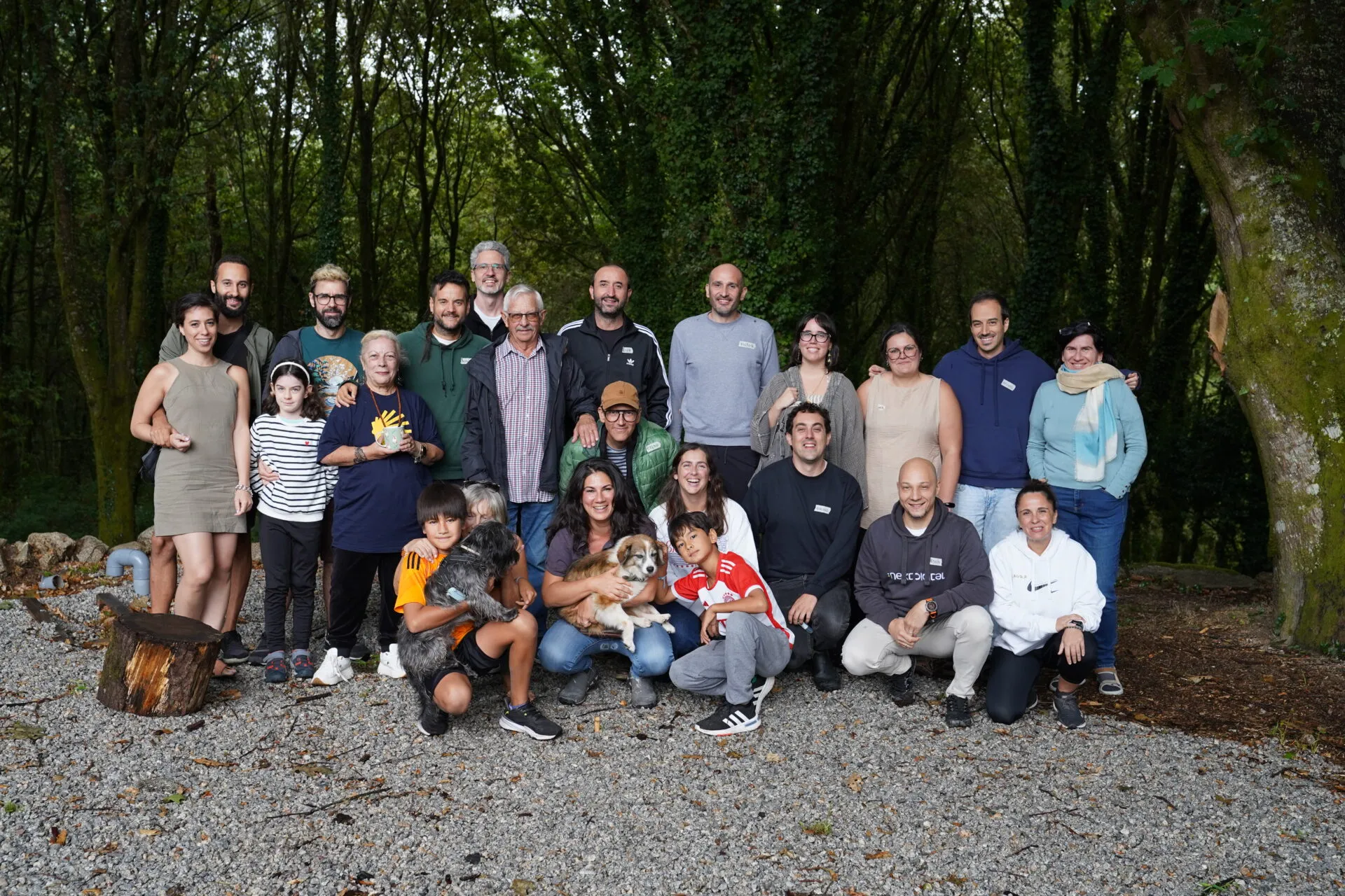 Grupo de unas veinticinco personas adultas y criaturas con dos perros posando sobre grava frente a un bosque verde y denso