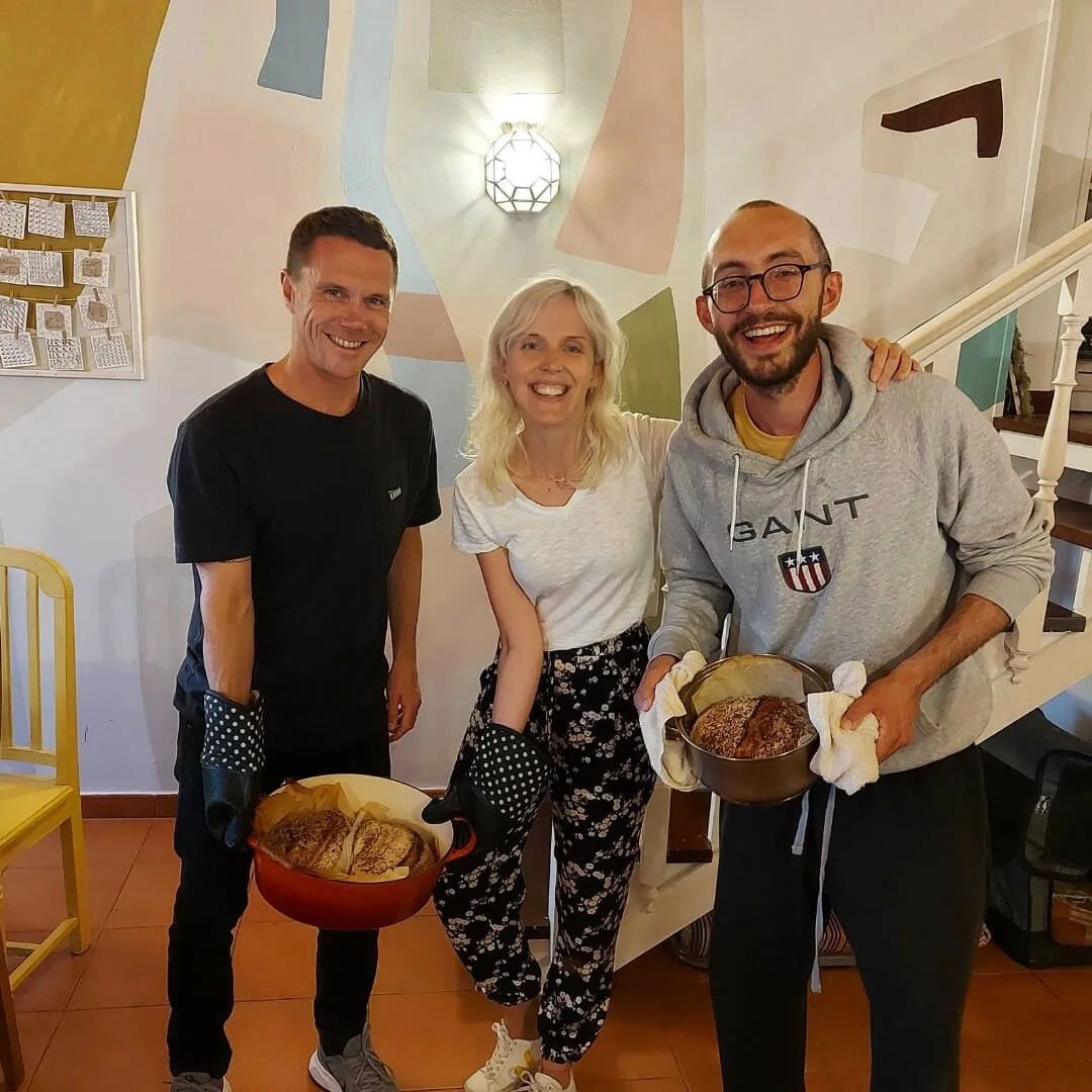 Tres colivers sonrientes sostienen cazuelas de hierro con pan recién horneado ante una pared con un mural en tonos pastel