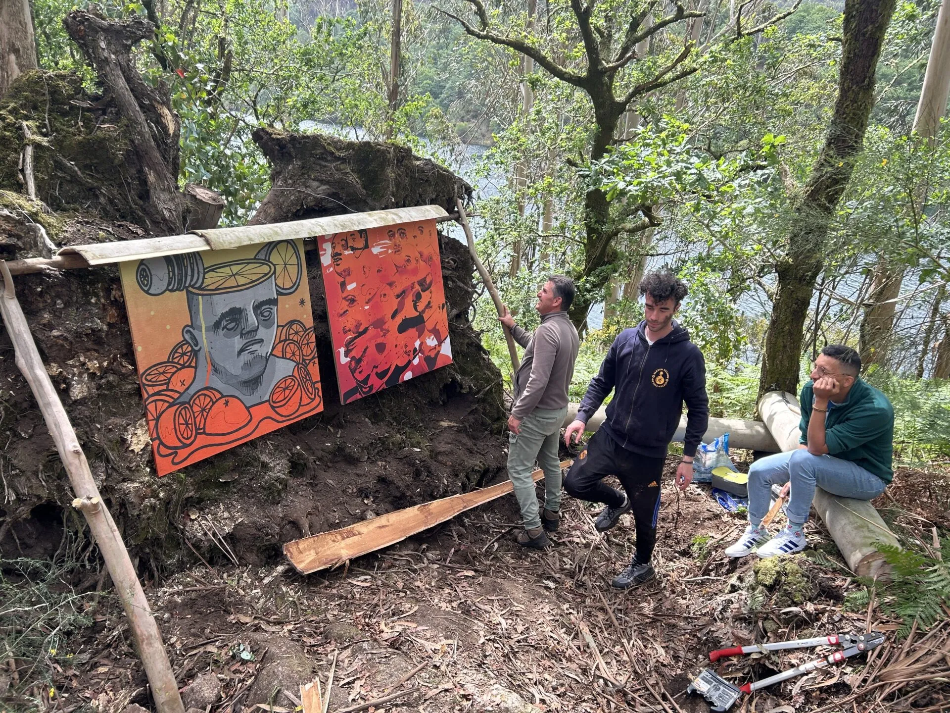 Tres homes nunha ladeira boscosa xunto a un río, colocando dous paneis pintados en tons laranxa sobre o tronco dunha árbore caída