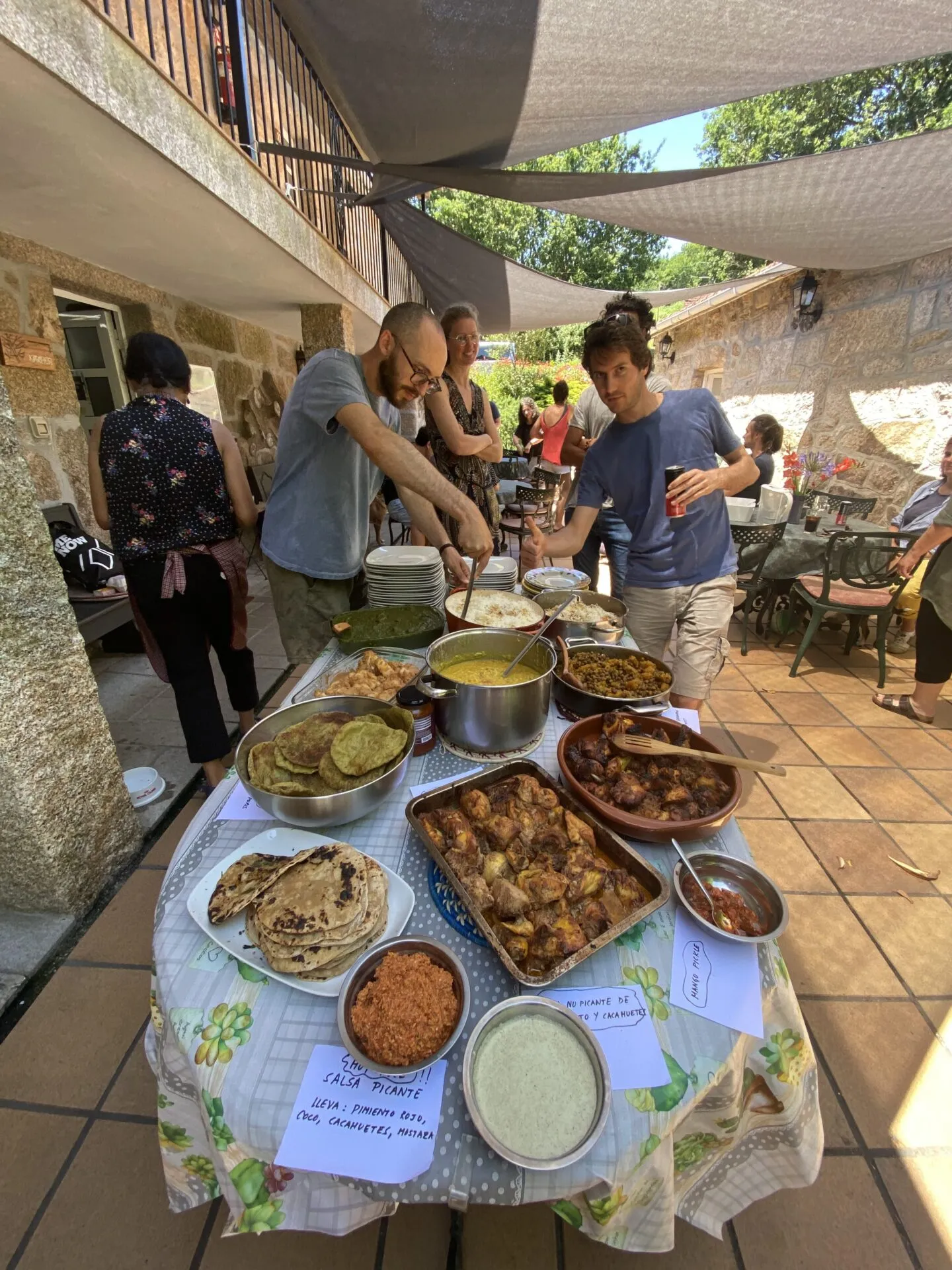 Mesa redonda nunha terraza sombreada chea de currys, arroz e pans planos, con carteis a man que din «salsa picante» e «mango pickle»