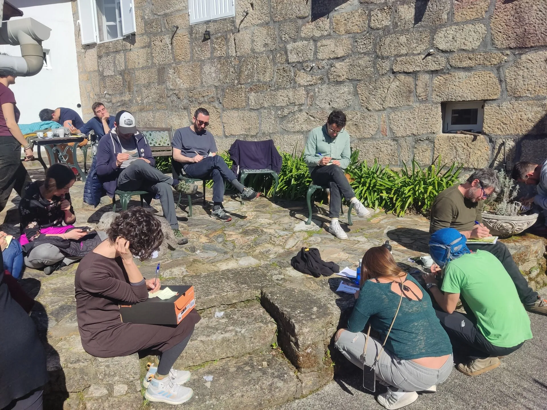 Una docena de personas repartidas por un patio de piedra soleado, escribiendo en cuadernos junto a un muro de granito