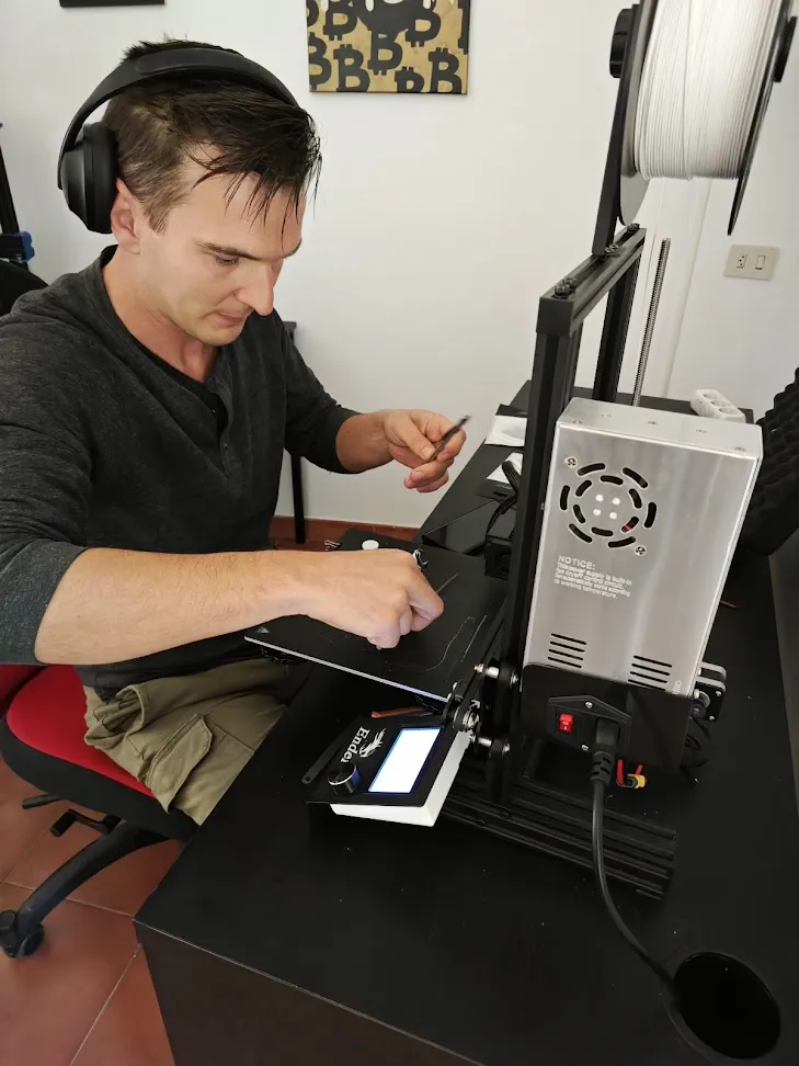 Home con auriculares traballando nunha impresora 3D de escritorio cunha bobina de filamento branco
