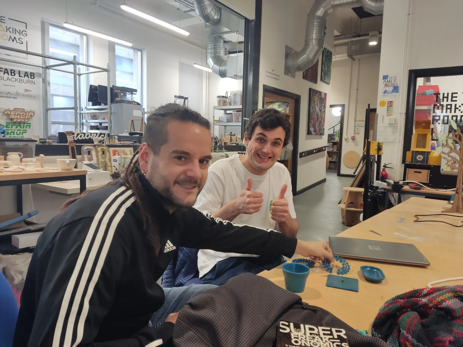 Dos hombres sonriendo en un banco de trabajo con piezas azules impresas en 3D en un taller con carteles de FAB LAB BLACKBURN y The Making Rooms