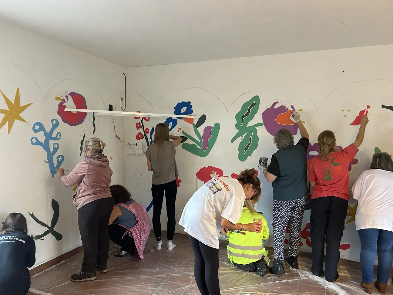 Arredor de oito mulleres pintando un mural floral de cores vivas nas paredes dunha sala branca, co chan cuberto de plástico