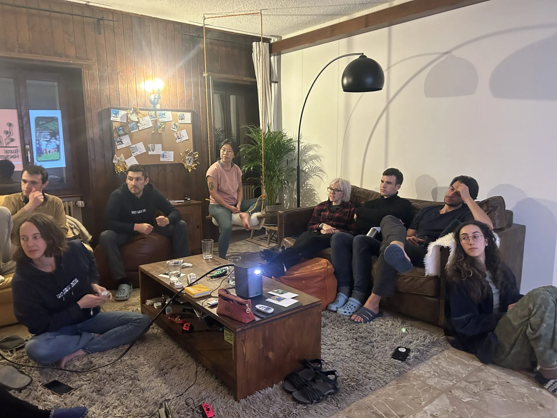 Nueve personas en sofás, pufs y el suelo de un salón con paredes de madera viendo una presentación proyectada