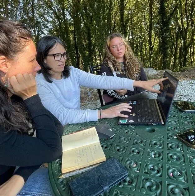 Tres mujeres alrededor de una mesa de jardín, una señalando la pantalla de un portátil junto a un cuaderno abierto