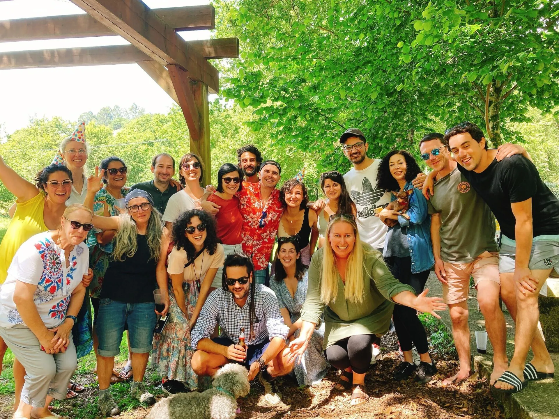 Around twenty cheerful people in summer clothes crowded together under a wooden pergola, with a small white dog at the bottom of the frame