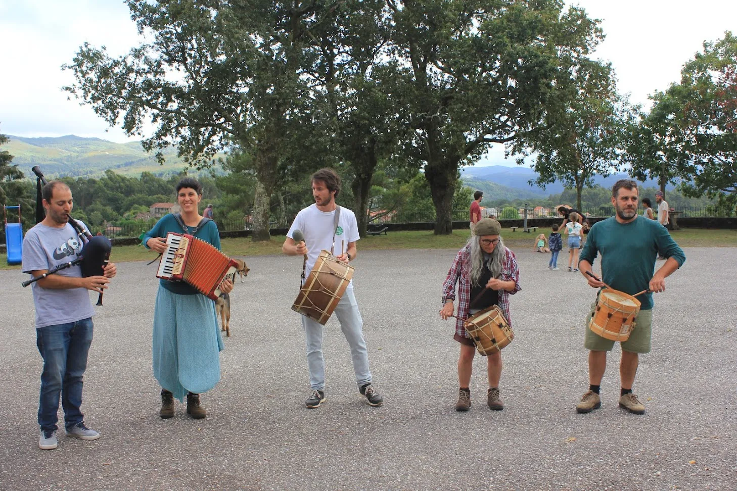 Cinco músicos tocan gaita, acordeón e tambores tradicionais nunha praza coas montañas ao fondo
