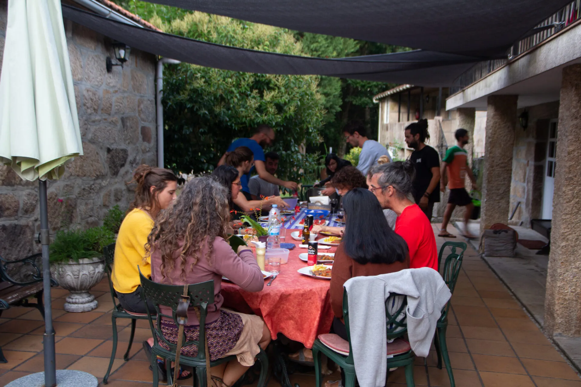 Unha ducia de persoas comendo xuntas nunha mesa longa baixo un toldo, na terraza de baldosa da casa de pedra