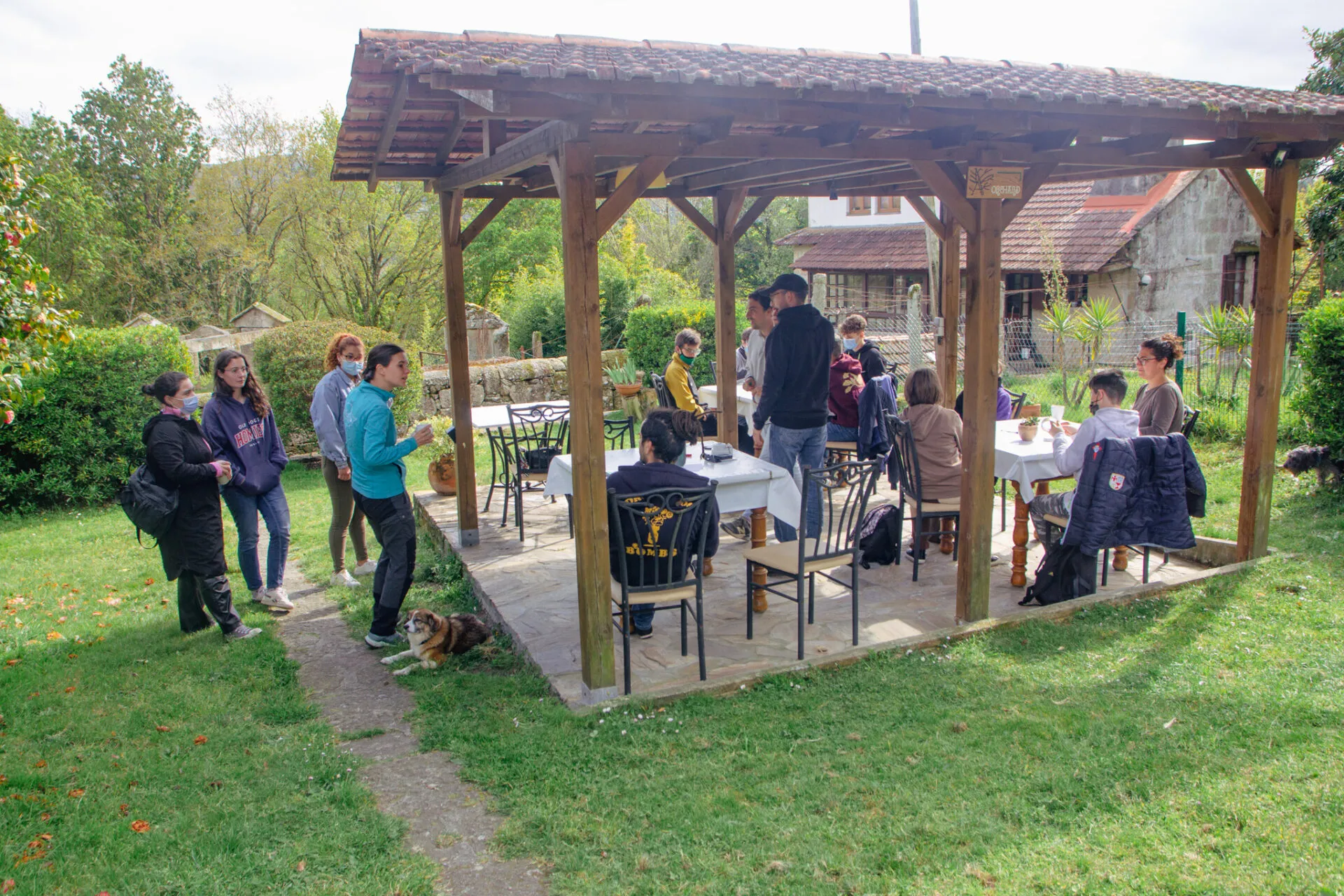 Personas con mascarilla reunidas alrededor de mesas bajo un cenador de madera con tejado de teja en un jardín verde