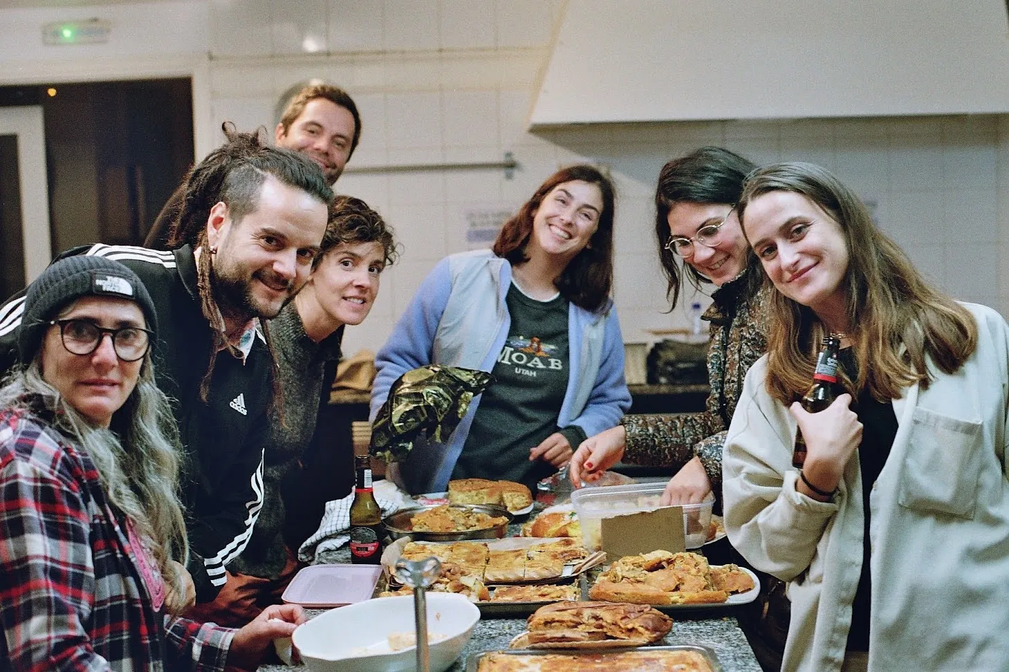 Seven people crowded round a kitchen counter loaded with trays of empanadas, smiling at the camera