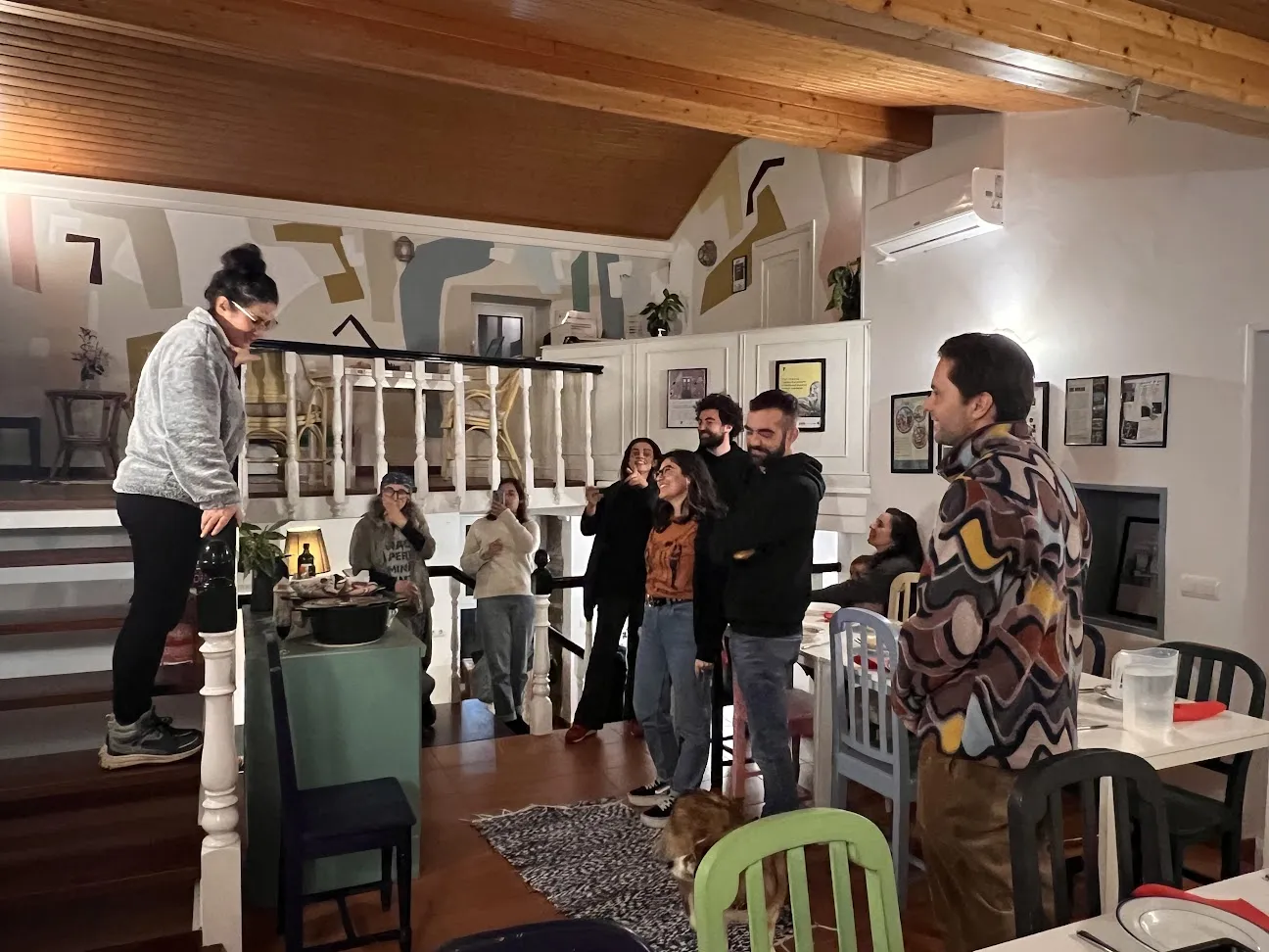 Mujer de pie sobre un escalón elevado hablando a un grupo que ríe en un comedor con mural pintado y techo de madera