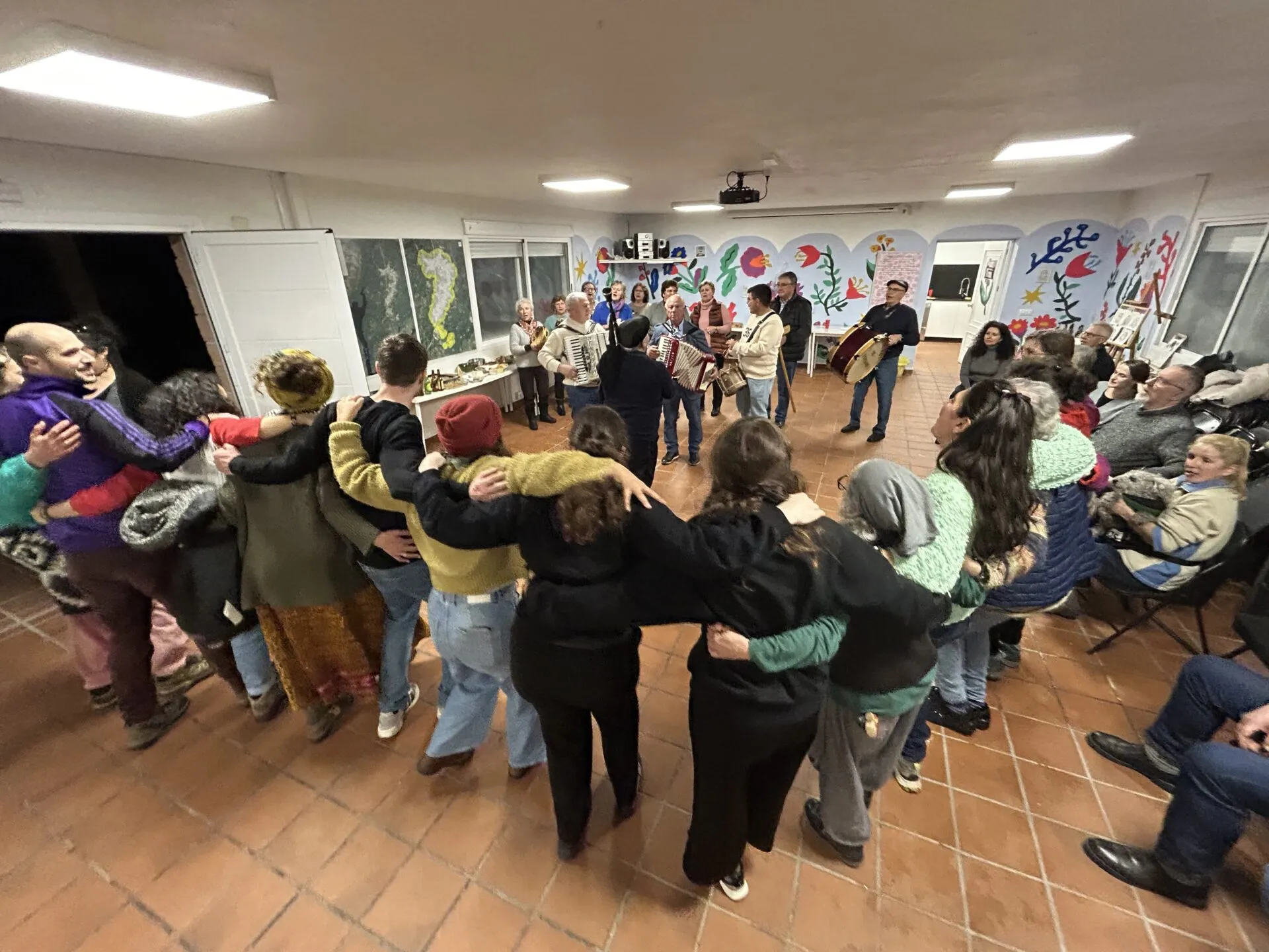 Un corro grande de xente bailando agarrada na Casa do Pobo, mentres a banda da aldea toca acordeóns e percusión ao fondo