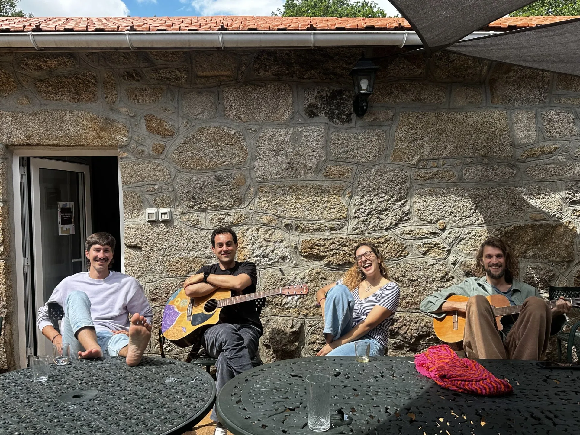 Catro persoas relaxadas contra un muro de granito nun patio, dúas delas tocando guitarras acústicas