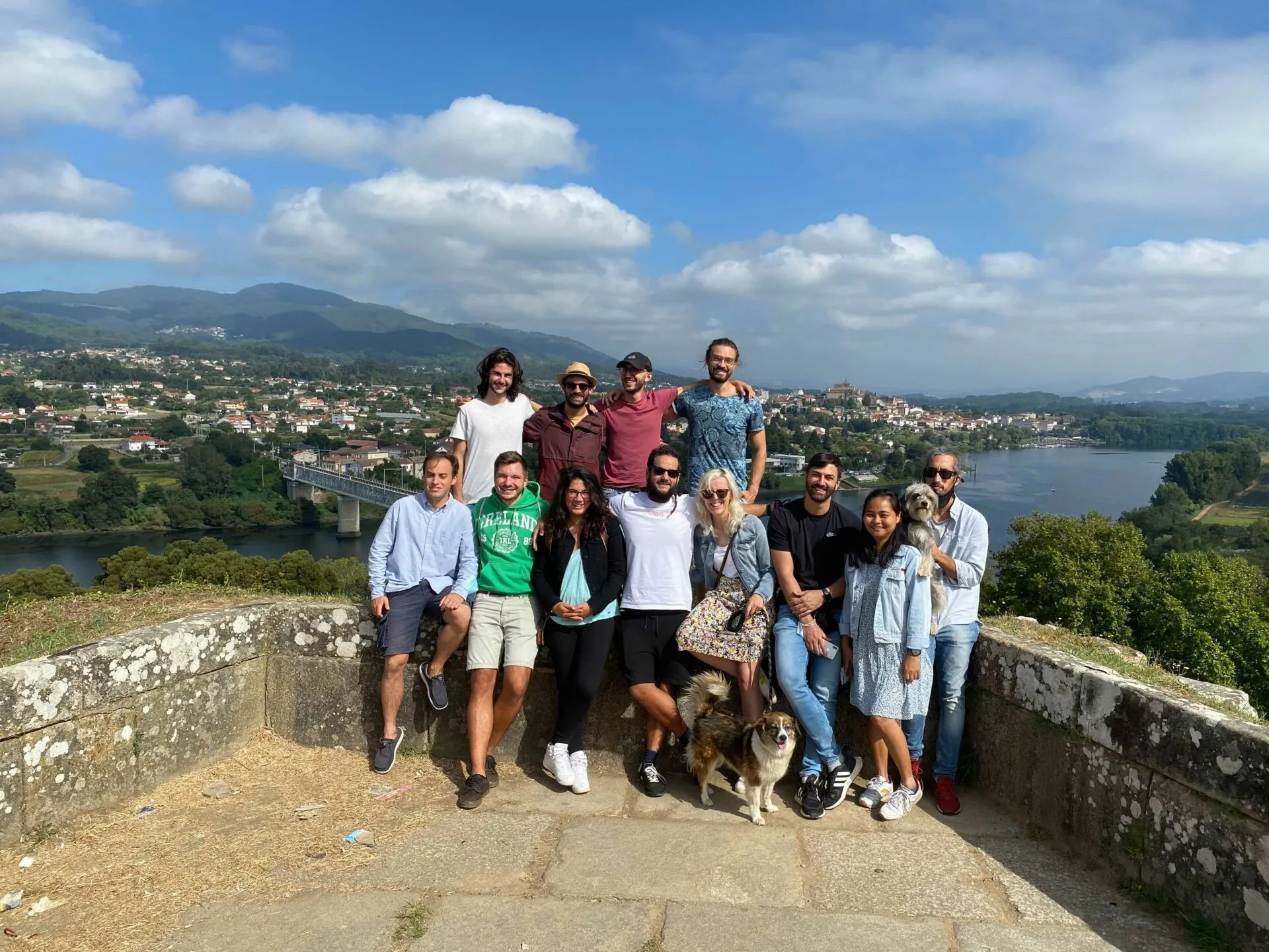 Once persoas e dous cans posando xunto ao muro dun miradoiro de pedra cun val fluvial e unha cidade detrás