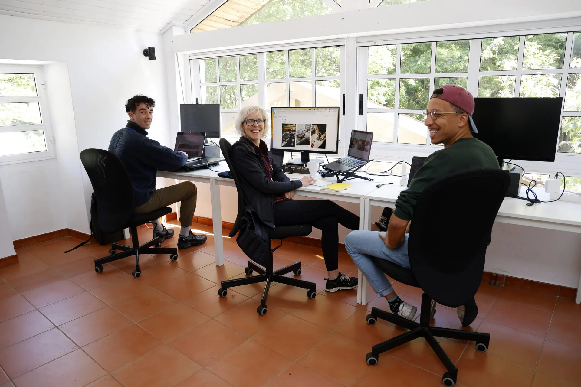 Tres colivers nas súas mesas do coworking acristalado, virándose cara á cámara e rindo, cos carballos ao outro lado das ventás