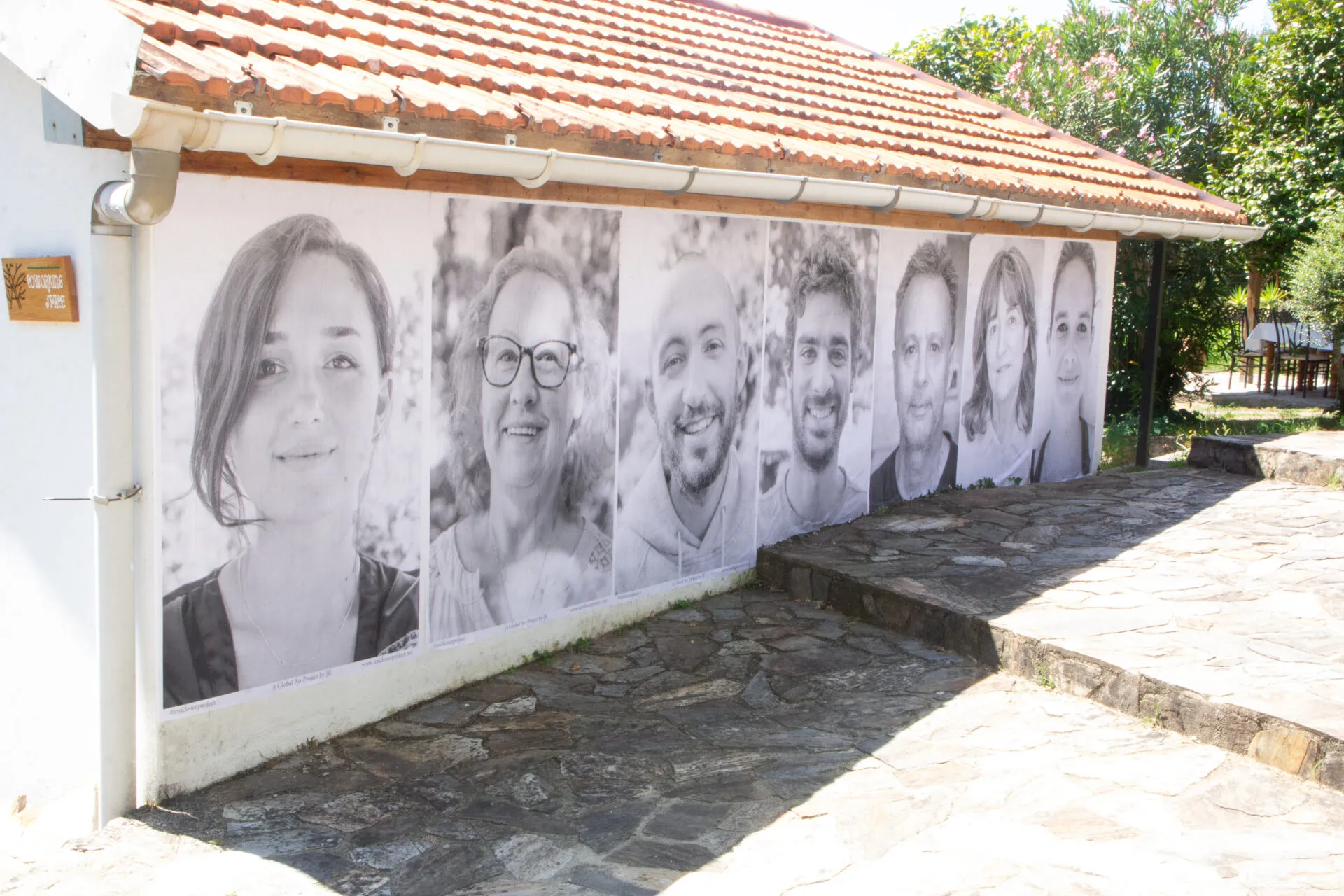 Unha fila de grandes retratos fotográficos en branco e negro pegados na parede exterior dun pequeno edificio de tellado de tella