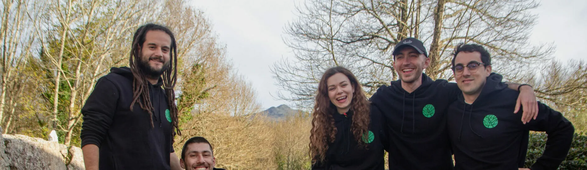 Recorte panorámico de cinco personas sonrientes con sudaderas negras con un pequeño logotipo verde, con árboles sin hojas detrás