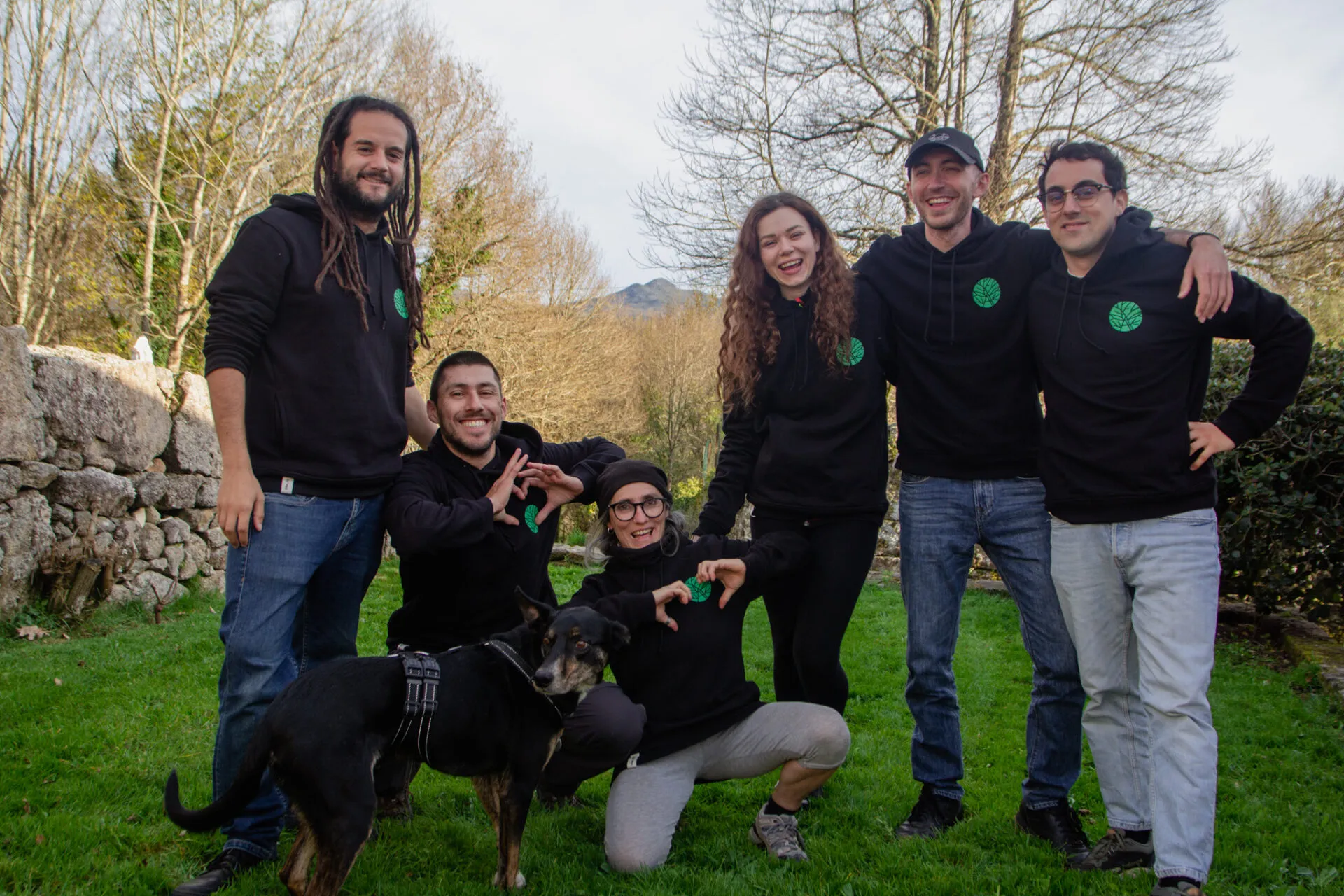 Seis personas con sudaderas negras iguales con un logo de árbol verde posando en la hierba con un perro negro