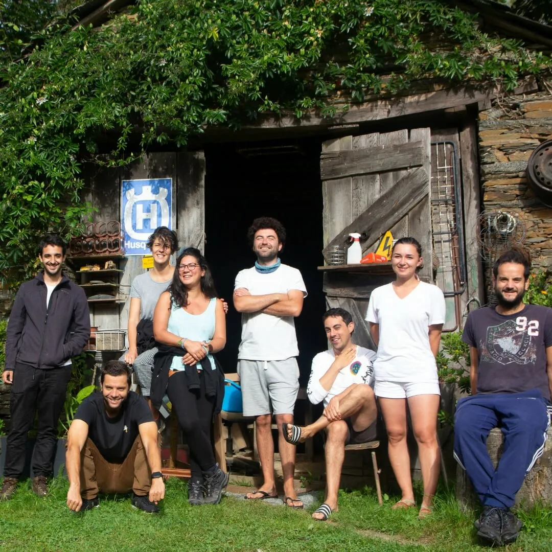 Nueve personas sonrientes posando en la hierba delante de un viejo galpón taller de piedra y madera