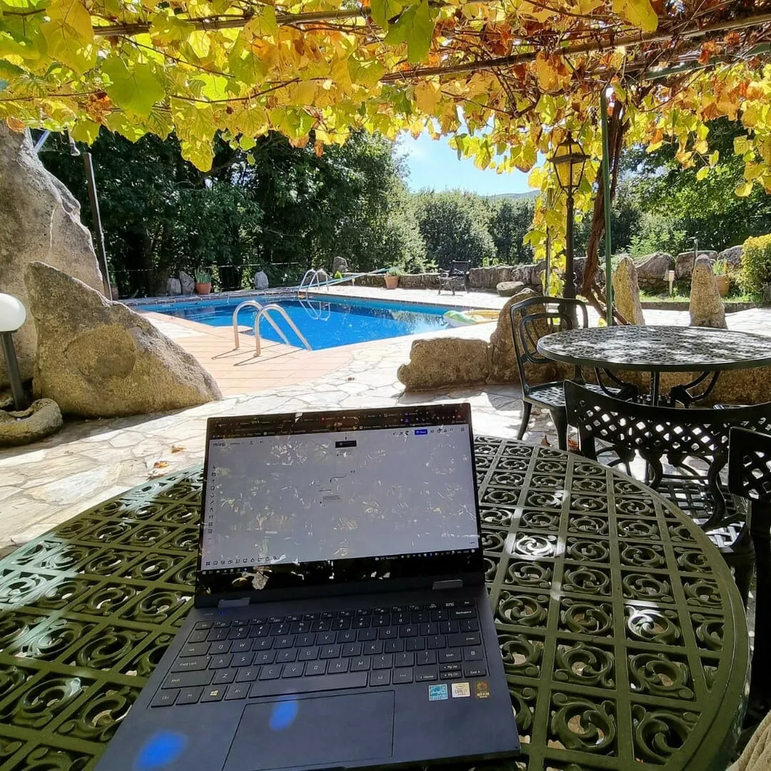 Portátil abierto sobre una mesa de jardín de metal verde bajo una parra, con una piscina azul al fondo