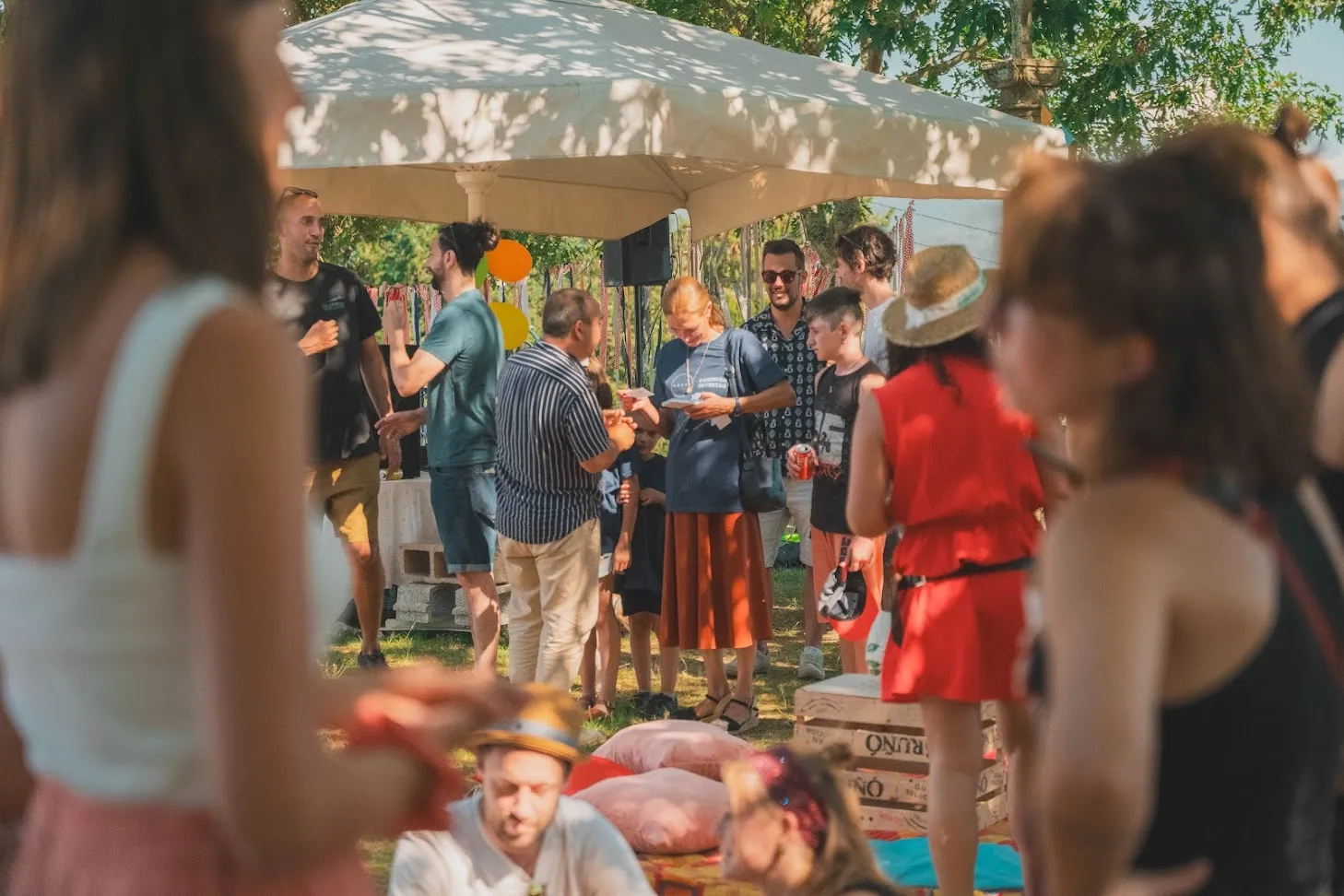 Convidados de pé conversando nunha festa ao aire libre baixo un toldo, con almofadas de picnic no chan