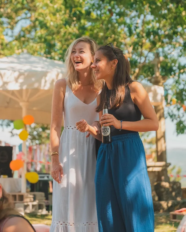 Dúas mulleres rindo nunha festa ao aire libre, unha con vestido branco e outra cunha cervexa, con bandeiroliñas e carpa detrás