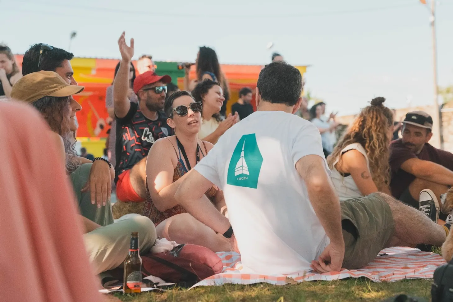 Xente sentada en mantas sobre a herba nun festival ao aire libre, conversando diante dun fondo pintado de cores