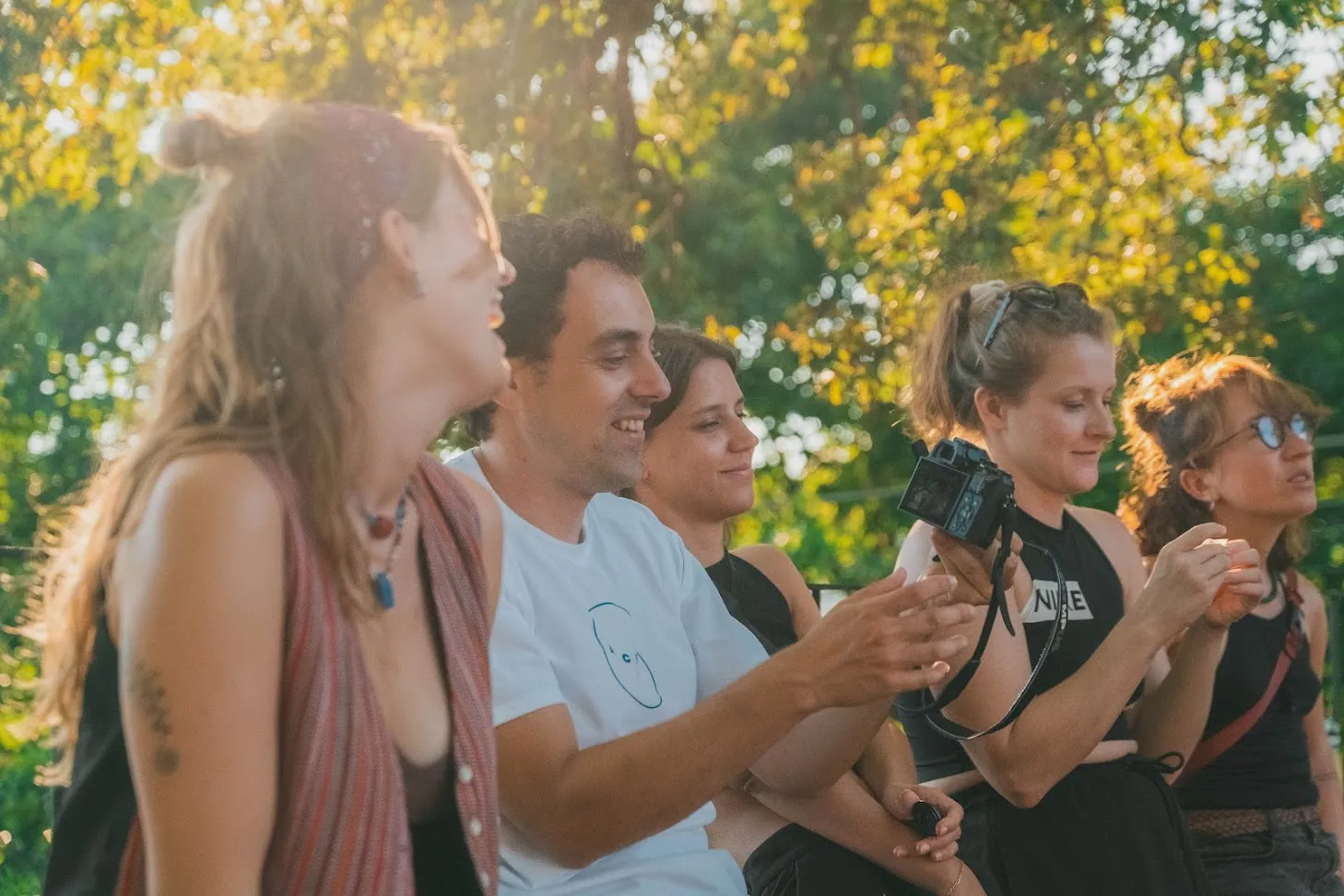 Cinco persoas sentadas en fila coa luz dourada do serán, rindo mentres unha sostén unha cámara