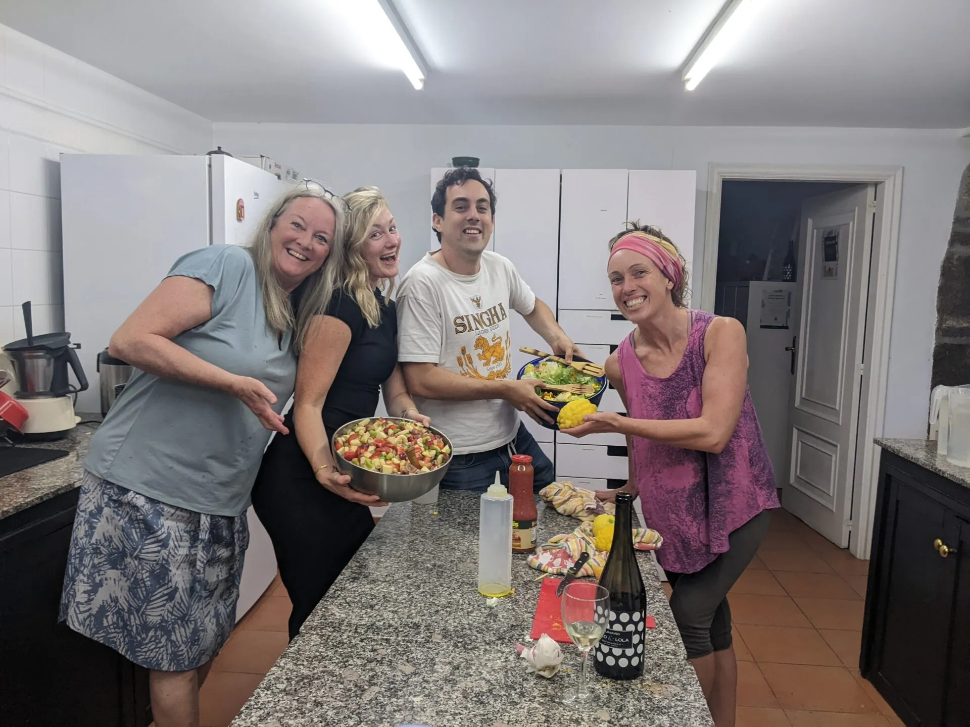Catro persoas rindo nunha cociña compartida, suxeitando grandes cuncas de ensalada e froita picada