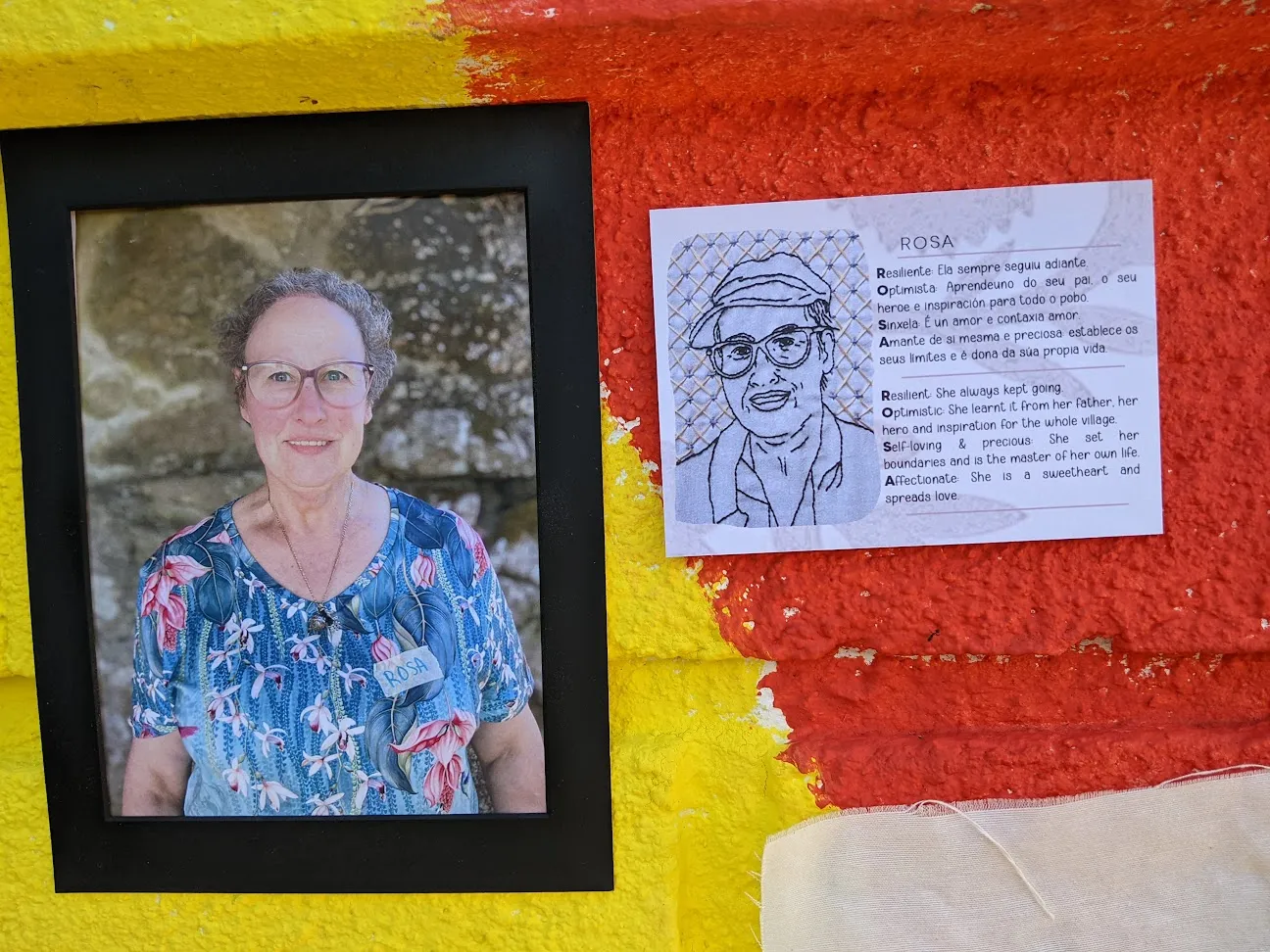 Retrato enmarcado dunha veciña a carón dun debuxo bordado e dun texto acróstico sobre Rosa, nun muro vermello e amarelo